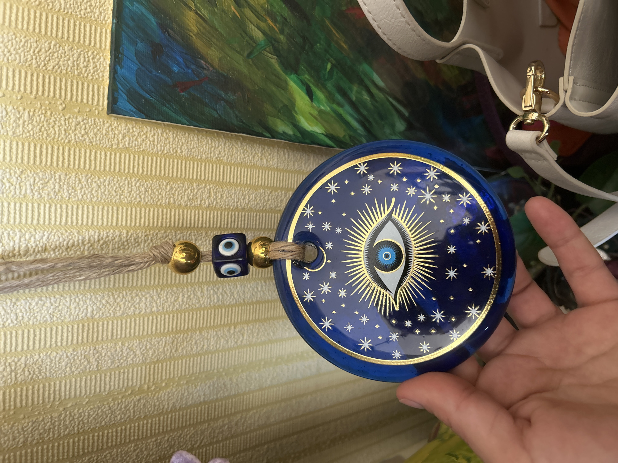 The image depicts a close-up of a decorative object held by a human hand, set against a background of a textured wall, artwork, and a handbag, within an indoor environment in the city of Unknown, Unknown.
The primary subject is a dark blue circular disc, approximately 15-20 centimeters in diameter, featuring a central "Evil Eye" motif. This motif consists of a white sclera, a light blue iris with a darker blue ring, and a central black pupil. Golden rays emanate radially from the eye. The disc's dark blue surface is further adorned with scattered golden stars of varying sizes and a small golden crescent moon in the upper left quadrant. Two concentric golden lines border the disc's circumference. A brown, twisted twine string passes through a hole at the top of the disc. Attached to this string, above the disc, are two spherical golden beads, flanking a single square dark blue bead. The square bead features two smaller "Evil Eye" symbols on its visible face.
A light-skinned human hand, with visible fingers, supports the bottom edge of the blue disc, presenting it towards the viewer. No jewelry or distinguishing marks are visible on the hand.
The background consists of several distinct elements. To the left and top, a light yellow wall covering, possibly wallpaper, exhibits a textured surface with both fine granular bumps and regular horizontal ribbed patterns. To the right of the central object, a section of a painting or canvas artwork is visible. This artwork features broad, visible brushstrokes predominantly in shades of green and blue, with hints of darker tones and possibly red. Below and to the right, a white leather or faux leather handbag is partially visible. Its upper rim, a white strap, and a prominent gold-colored swivel snap hook clasp are discernible. An obscured interior suggests an orange or brown lining or contents within the bag.