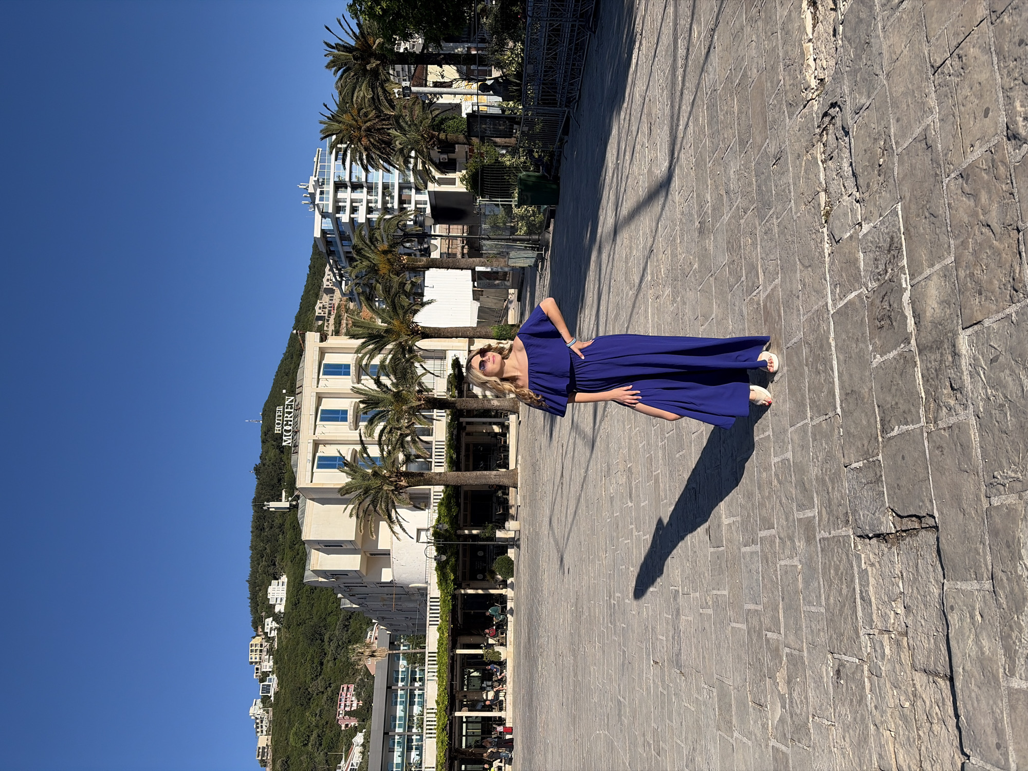 A woman in a long, flowing blue dress stands on a paved walkway in Budva, Montenegro. The background includes palm trees and buildings, with "Hotel Mogren" visible on one structure. The sky is clear and blue. People are seated at tables visible in the background. The woman is posing with her hand on her hip.