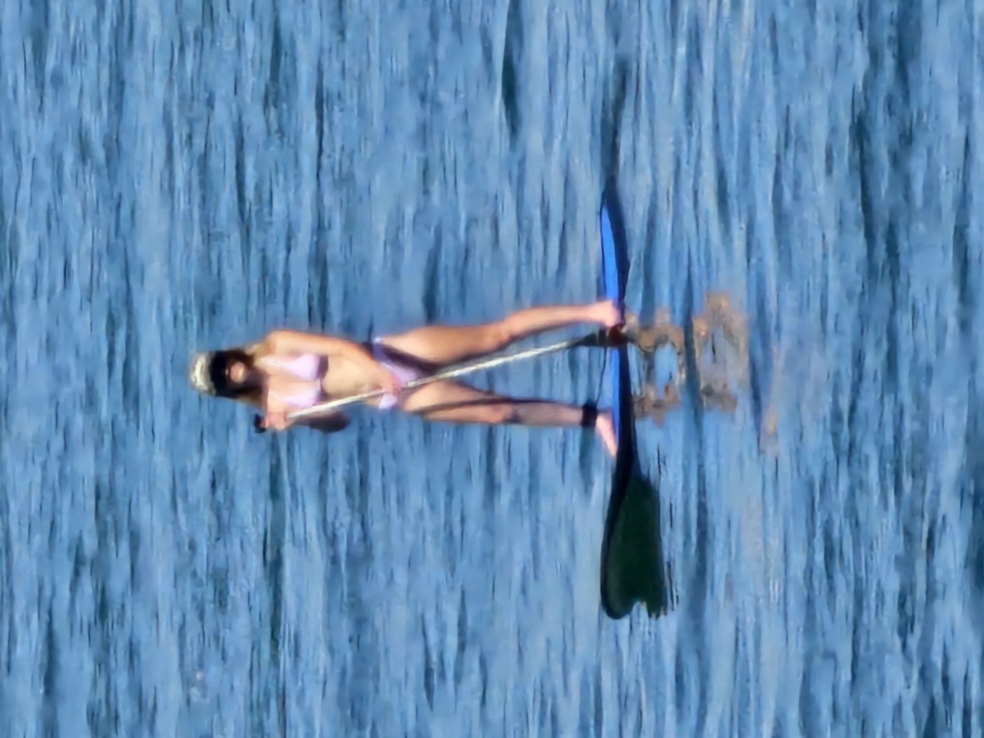 In the open water off the coast of Denpasar, Indonesia, a person stands on a paddleboard. The individual is wearing a light pink bikini and a camouflage-patterned baseball cap, with sunglasses obscuring their eyes. They are holding a paddle, with the blade submerged in the water, suggesting they are actively paddling. The paddleboard is blue and appears to be moving through the blue, rippling water, with reflections visible below. The person is tethered to the board with a black ankle leash. The overall scene captures a moment of recreational activity on the water.