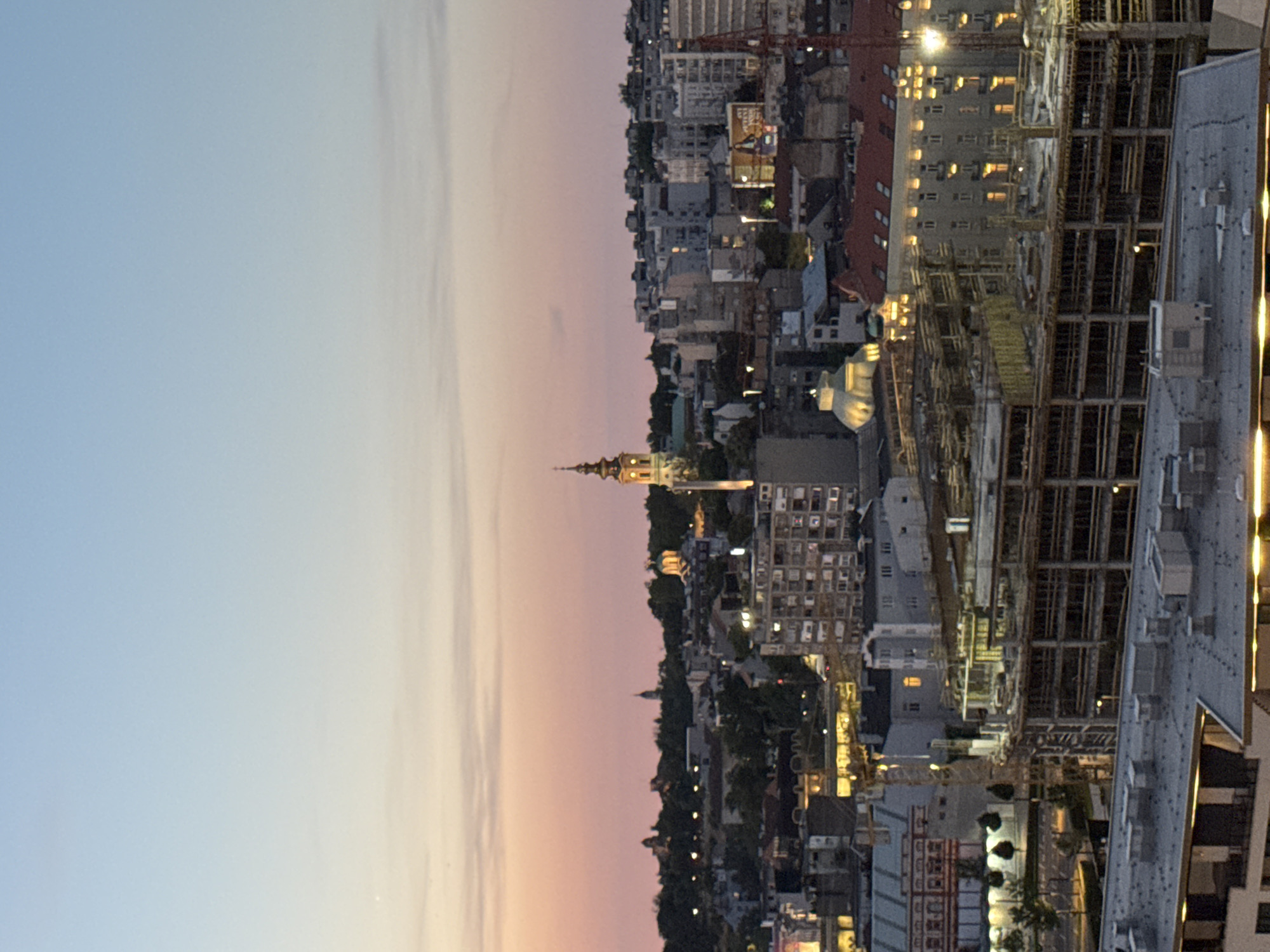 This image shows a skyline in Belgrade, Serbia, at dusk. The sky transitions from light blue to shades of pink and orange near the horizon. The city is dense with buildings, including residential structures and structures under construction, marked by visible cranes. A prominent church tower, lit with warm light, stands out among the urban landscape. Artificial lights illuminate windows and streets, creating a contrast with the fading daylight. The vantage point is high, likely from an elevated position within the city.