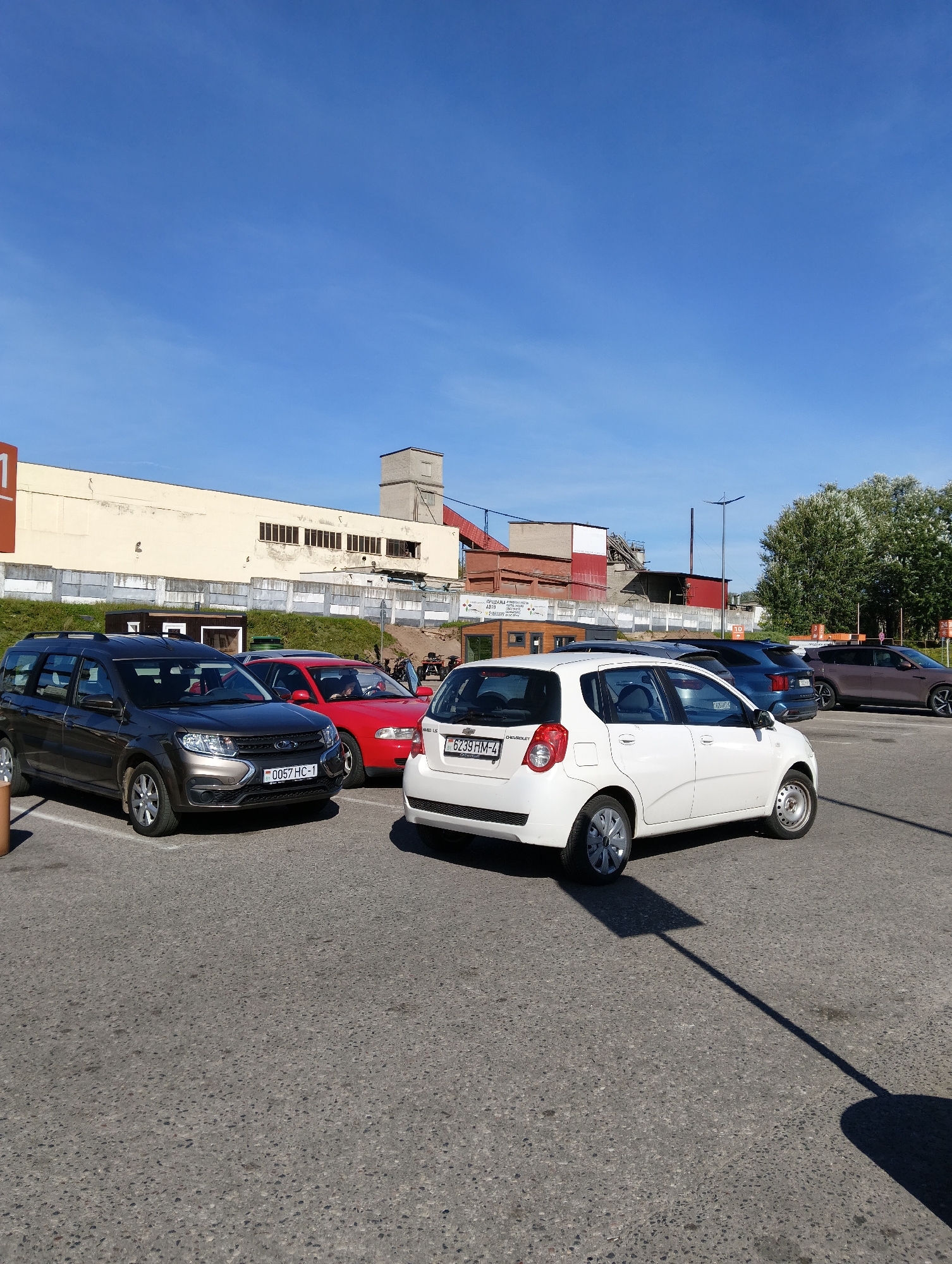 This is a photograph of a parking lot in Hrodna, Belarus. A dark gray Lada Vesta wagon is parked in the foreground on the left, with its license plate reading "0057 HC-1". To its right is a red sedan, followed by a white Chevrolet Aveo hatchback. In the background, there is a long, cream-colored building with windows, possibly a factory or warehouse, and further behind, several trees. The sky is clear and blue.