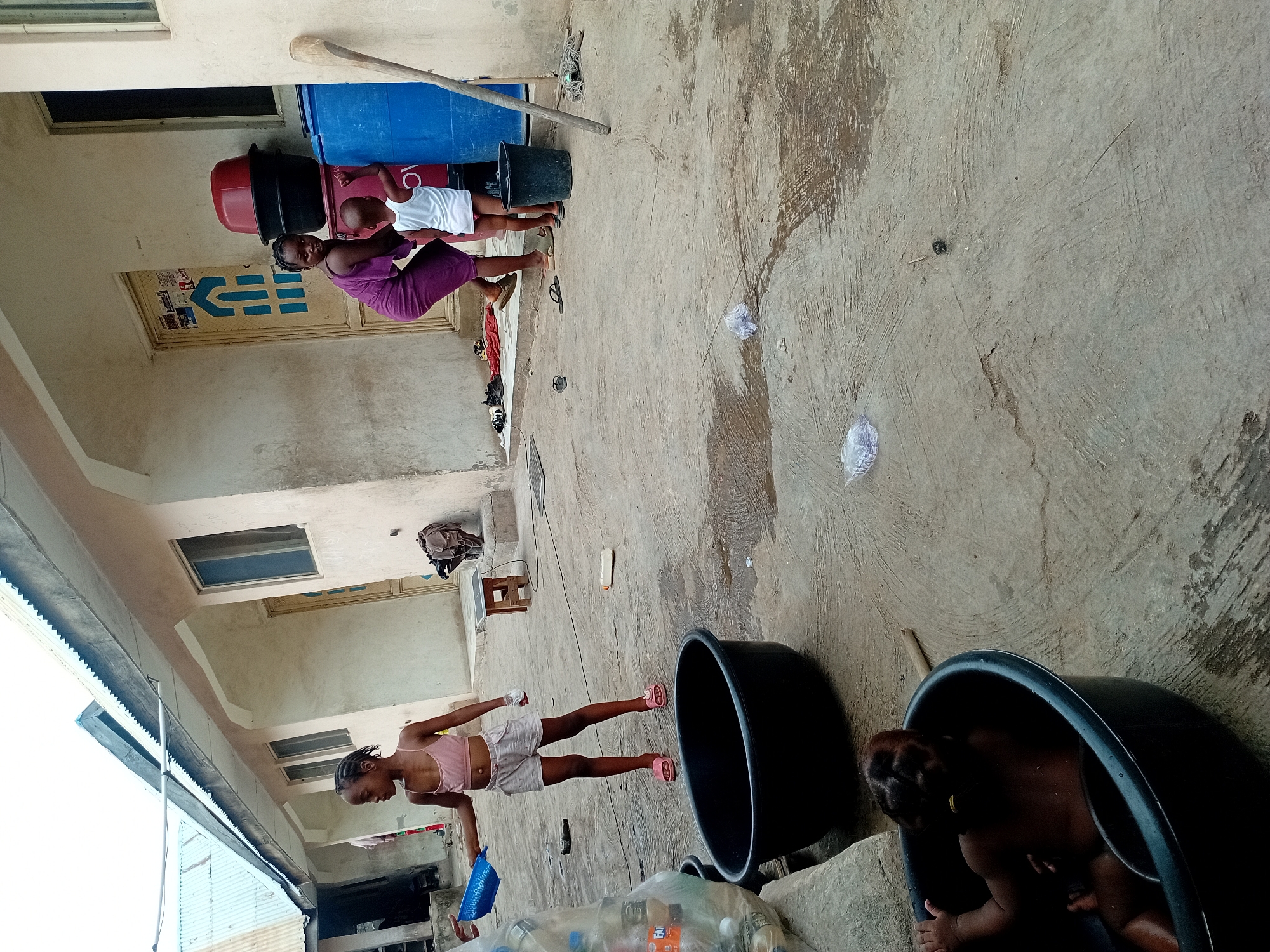 The image shows an outdoor scene in Unknown, Unknown, possibly a courtyard or backyard.
A woman in a purple dress stands next to a red and black object, attending to a child in white clothing. A blue barrel leans against the wall behind them.
In the foreground, a girl in a pink top and white shorts stands near a collection of plastic bottles and a blue tray. Two black tubs are visible; one appears empty, and the other contains a child. The overall environment appears to be a residential area with simple construction.