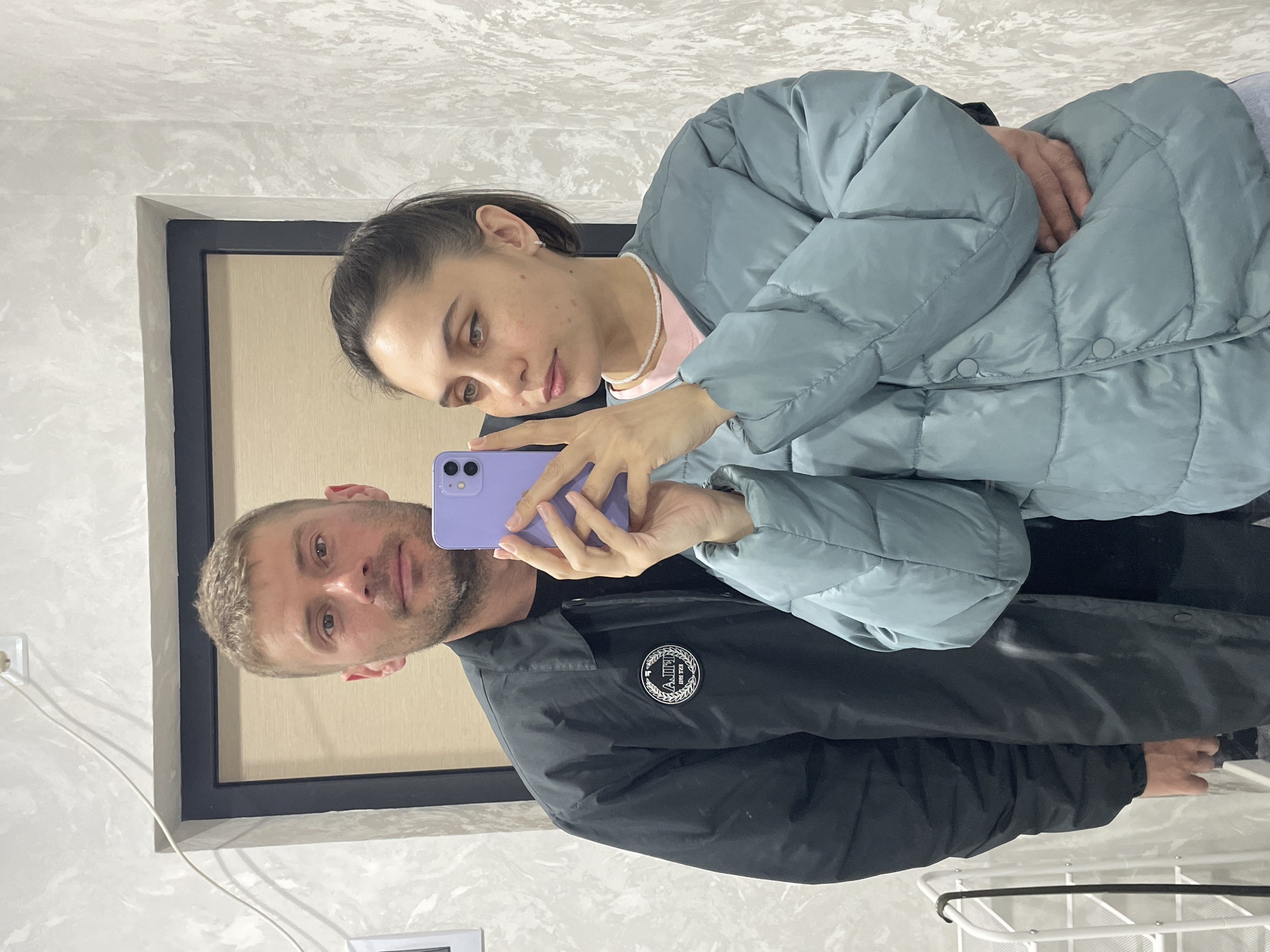 A man and a woman are taking a selfie in a room. The man has short, light hair and is wearing a black jacket with a circular emblem on the left breast. The woman has her hair pulled back and is wearing a light blue puffer jacket over a pink shirt with a pearl necklace. She is holding a purple smartphone. The room has a light-colored wall with textured patterns, an electrical outlet, and a white rack in the bottom right corner. There is a window or a framed opening behind them. No details indicate that the location is Izhevsk, Russia.