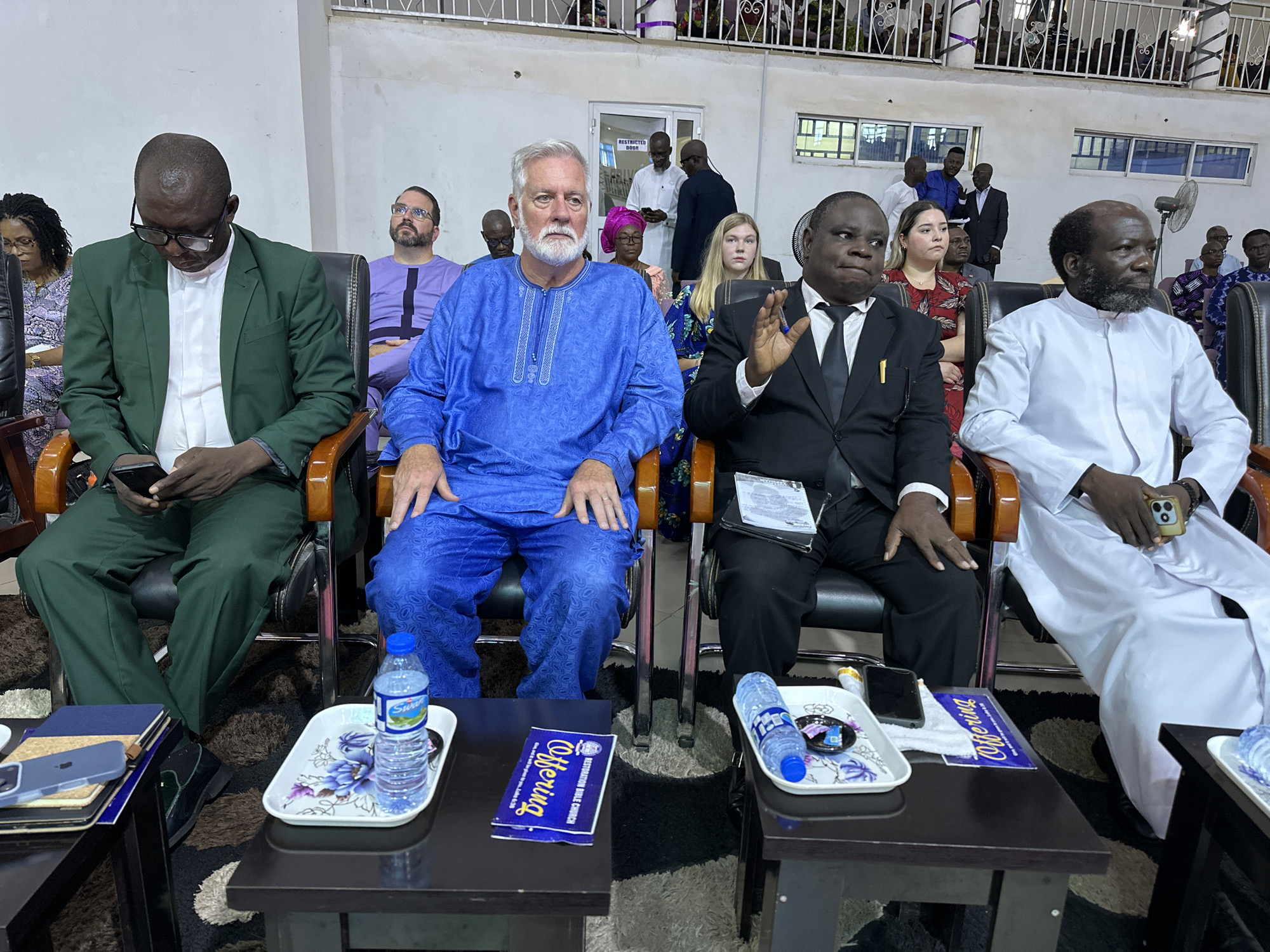 This image captures an indoor gathering, likely a religious service, taking place in Television, Nigeria. The foreground is dominated by several individuals seated in dark chairs. A man in a green suit and glasses looks down at his phone. Beside him, a man in a vibrant blue garment with intricate patterns sits attentively. To his right, a man in a black suit and tie gestures with his hand. Further to the right, a man in a white robe is seated. In front of the attendees are dark wooden tables holding various items, including books, smartphones, offering envelopes, and bottles of water. The background reveals more people seated in rows, some standing near a door labeled "RESTRICTED DOOR," and a white ceiling with a railing. The overall atmosphere appears formal and contemplative.