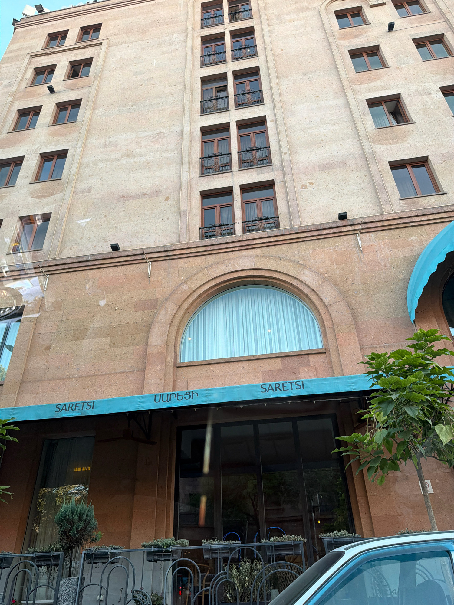 The image shows a low-angle exterior view of a multi-story building with a facade of textured, reddish-brown stone. The building has numerous windows with wooden frames, many of which appear to be open or have balconies. A light blue awning extends across the lower section of the building, bearing the text "SARETSI" in white capital letters, along with text in Armenian script. Below the awning, large glass windows reveal the interior of what appears to be a commercial establishment. To the right of the awning, a tree with green foliage grows. In the foreground, the hood and windshield of a light-colored car are visible. The overall scene suggests a street-level view in an urban environment. The text "SARETSI" suggests this is likely a commercial establishment, possibly a restaurant or shop, located in Yerevan, Armenia.