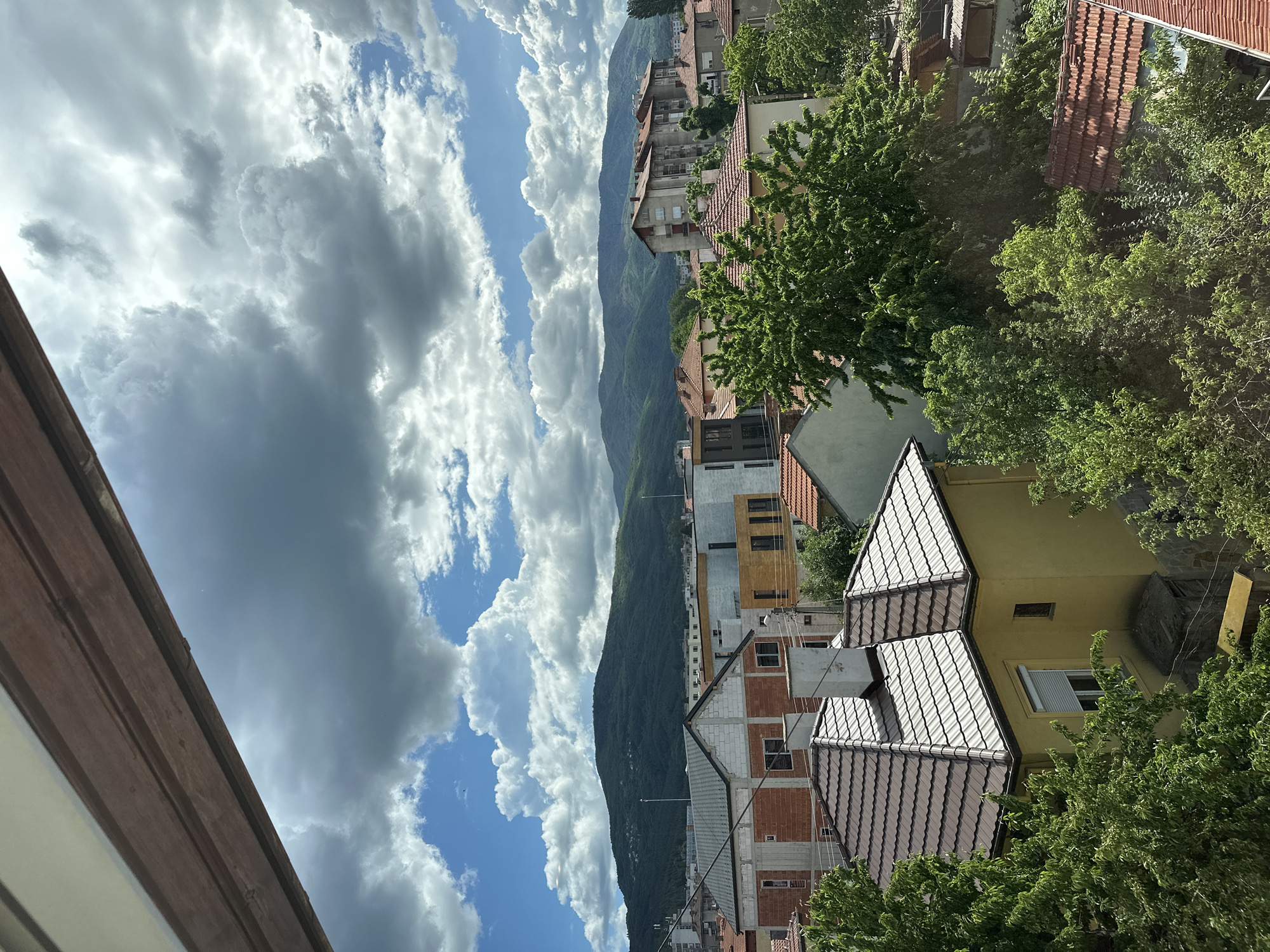 The image depicts a cityscape with a mountain range in the background under a cloudy sky. In the foreground are buildings with tiled roofs, some surrounded by trees. The architecture varies, with some structures appearing newly constructed with exposed brickwork, while others seem older and painted. The sky is filled with a mix of dark, stormy clouds and patches of blue sky. The camera angle is slightly tilted, providing a view from an elevated position.