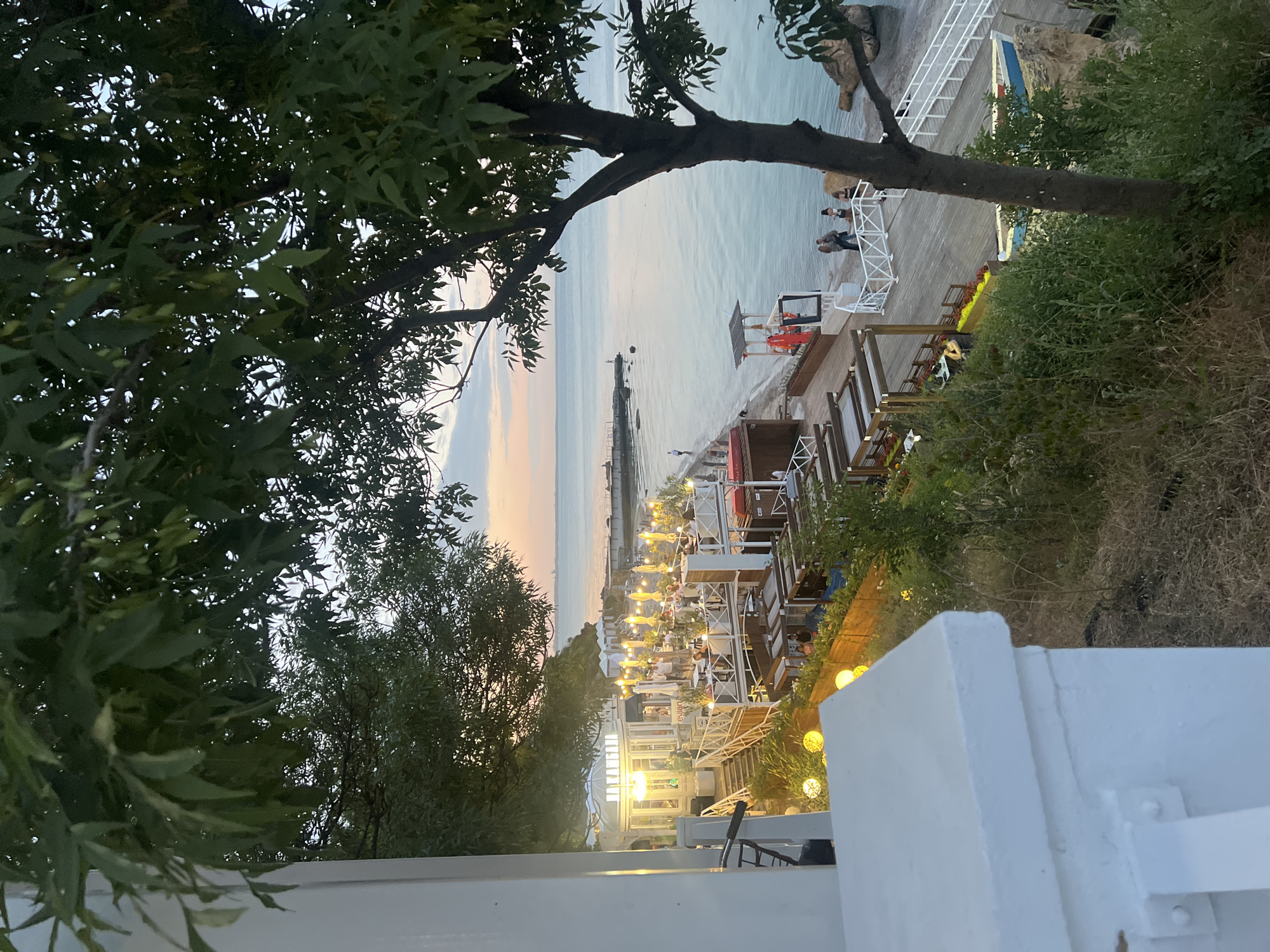 An outdoor dining area is situated on a raised wooden platform overlooking the Black Sea in Odesa, Ukraine. The area is illuminated by numerous string lights and features multiple tables with people seated. The scene is framed by lush green trees, with a large tree on the right side of the image. In the background, the sea stretches to the horizon, with a hint of the Odesa cityscape visible in the distance. The sky displays the soft hues of either sunrise or sunset. A lifeguard stand is visible on the beach below, with a few individuals walking along the shore. The overall atmosphere suggests a relaxed evening dining experience by the sea.