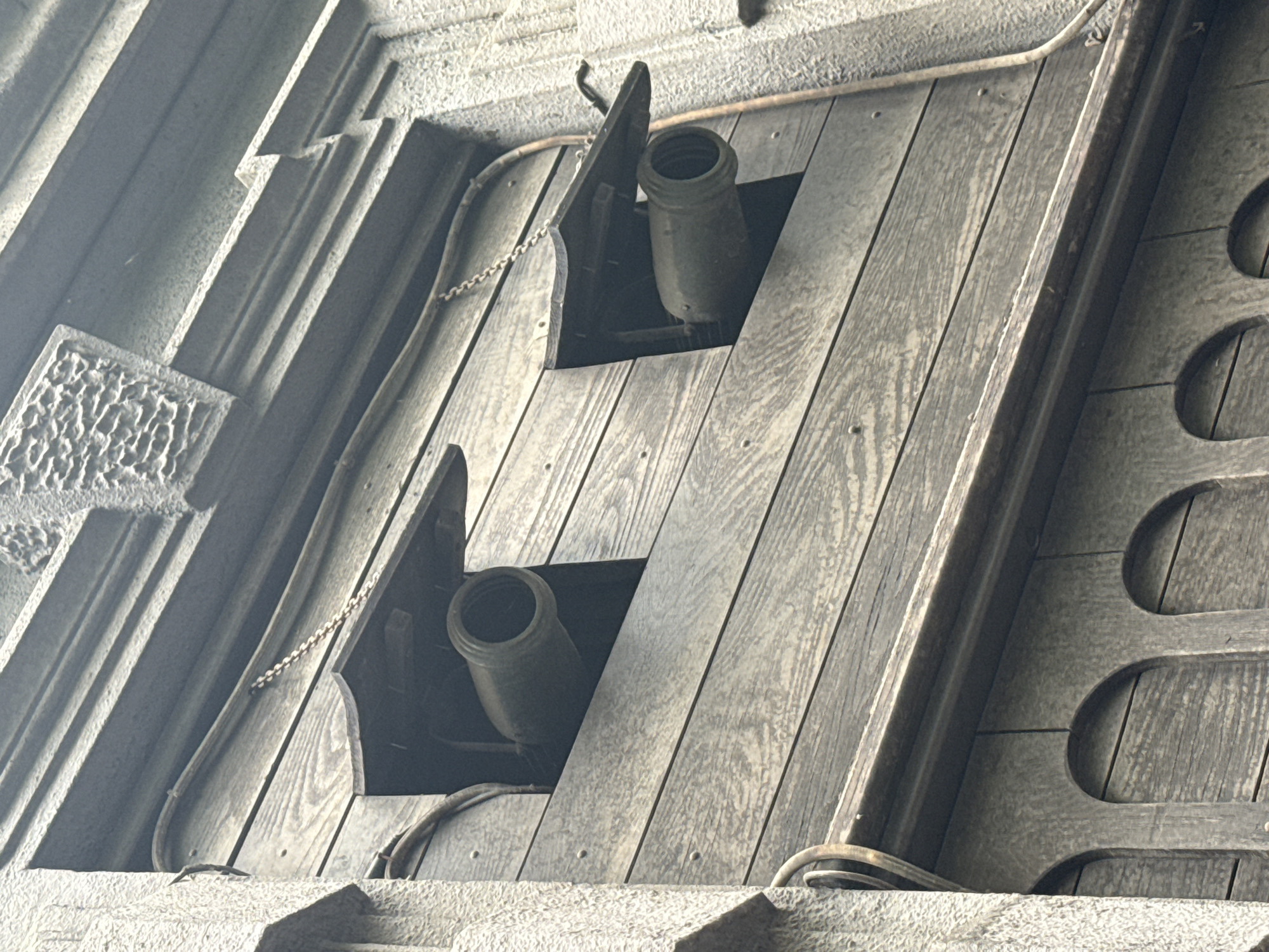 The image depicts two cannons positioned behind wooden shutters in Zagreb, Croatia. The cannons are a dark, metallic color, possibly a weathered gray or green, and are aimed downwards from their respective openings. The shutters are constructed of light-colored wood planks arranged vertically. The overall environment appears to be part of an old stone or concrete structure, with decorative molding visible on the left side of the image. The lighting suggests it is daytime, but there is some haze or mist in the air, creating a slightly muted effect.