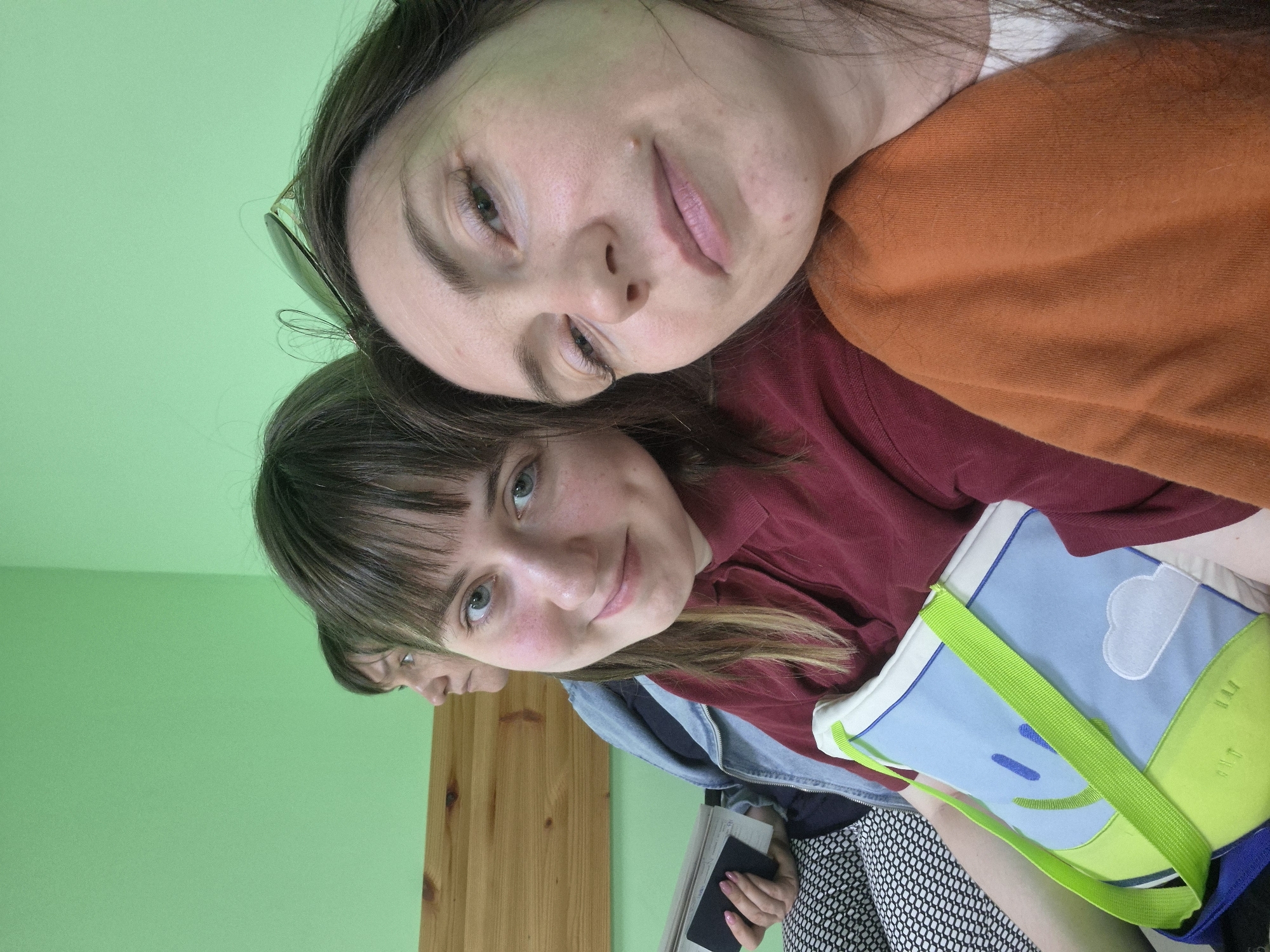Three females are in a room with green walls and a wooden door or panel. Two are prominently visible: one with brown hair and bangs, wearing a burgundy shirt and a denim jacket, holding a notebook or tablet, and another with long brown hair and sunglasses atop her head, wearing an orange sweater. The third female is partially visible in the background, near the door/panel. The foreground female holds a blue and green bag with a cloud design. The image appears to be a selfie or taken at close range. The city and specific location are unknown.