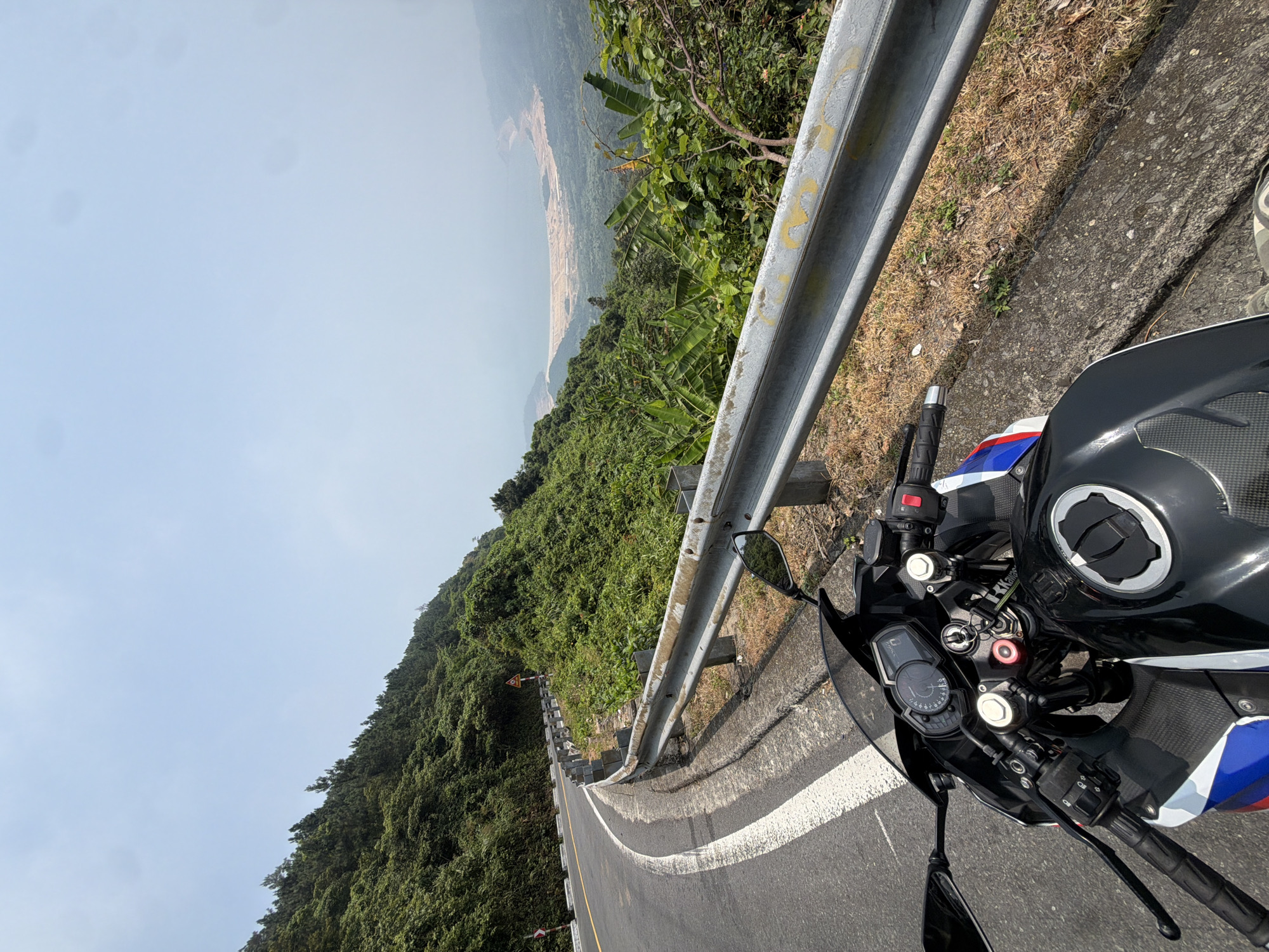 A motorcycle is parked on the side of a winding mountain road. The road has a white line marking the edge and a metal guardrail. Lush green vegetation covers the mountainside. In the distance, there is a hazy view of the coastline. This road is likely on or near Son Tra Mountain, also known as Monkey Mountain, a popular destination in Đà Nẵng, Vietnam. The motorcycle appears to be a modern sportbike, with its handlebars, gauges, and fuel tank visible.