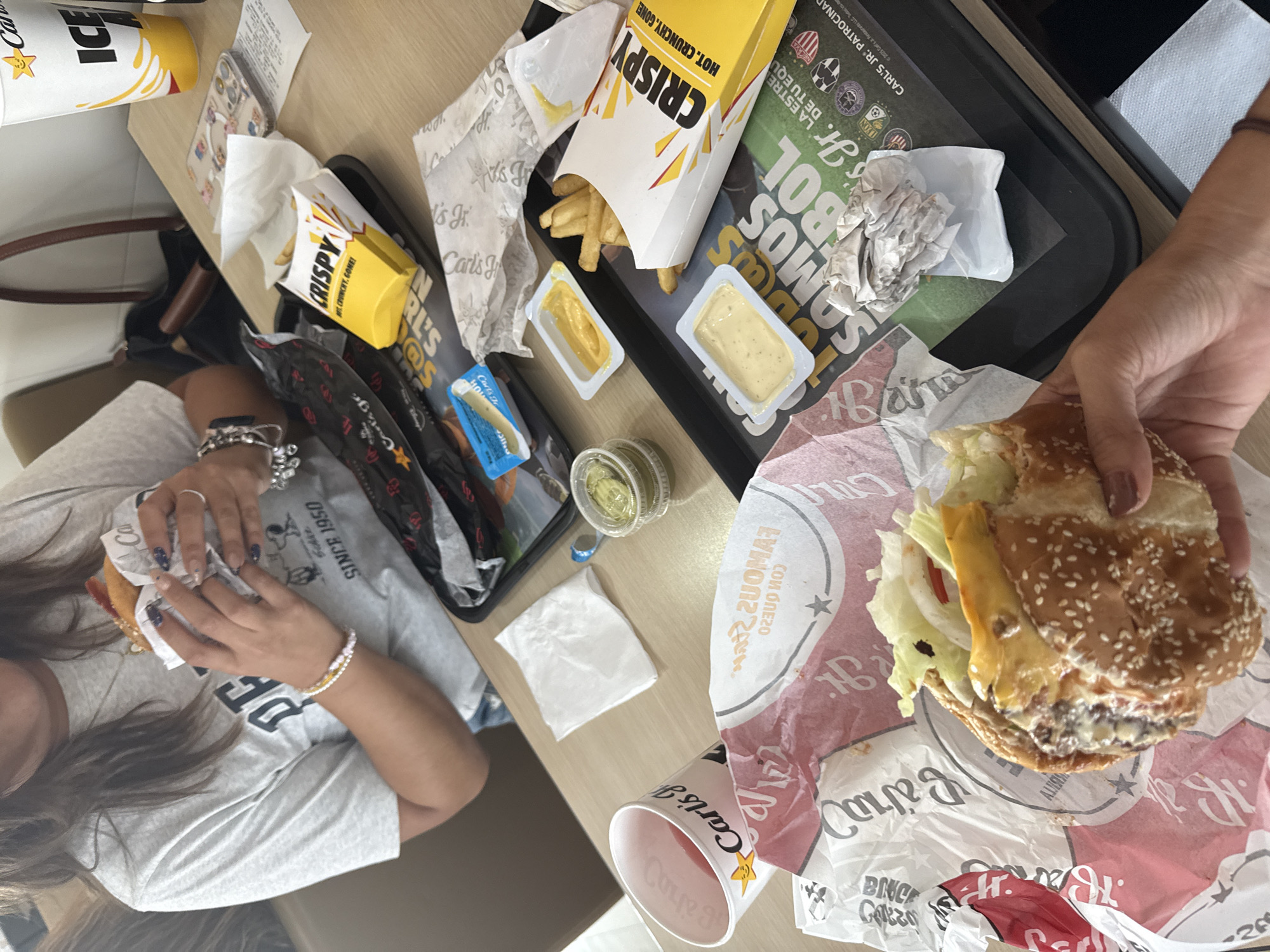 Here's a description of the image:
The image shows an overhead view of a table at what appears to be a Carl's Jr. restaurant. Two people are visible, each holding a burger. One person is on the left side of the frame, and the other is on the right.
On the table, there is a black tray with several Carl's Jr. items, including a box of "CRISPY" fries, sauce containers, and crumpled paper wrappers. There's also a cup with the Carl's Jr. logo on the table. The food suggests a meal at a fast-food restaurant.
Given the presence of Spanish writing on the tray, and the prompt mentioning San Bernardino Tlaxcalancingo, Mexico, it can be assumed that the location is a Carl's Jr. branch in that city.