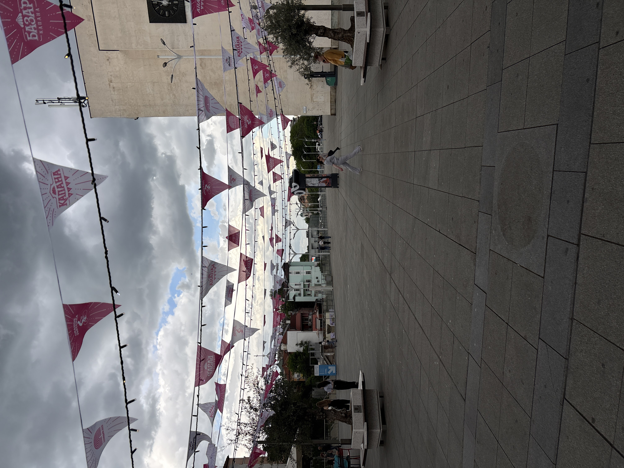 The image captures a street scene in Plovdiv, Bulgaria, identified by the "Kapana Bazaar" flags strung overhead. The location is a wide, pedestrianized area paved with gray stone blocks. A person is captured mid-leap in the center of the walkway, suggesting movement or play. Other individuals are visible in the background, engaged in various activities such as standing, walking, or sitting on benches. Trees and benches are positioned along the sides of the walkway. Buildings line the street in the distance, with one building featuring a clock on its facade. Above, string lights with triangular pennants are suspended across the street. The sky is overcast with a mix of clouds and patches of blue.