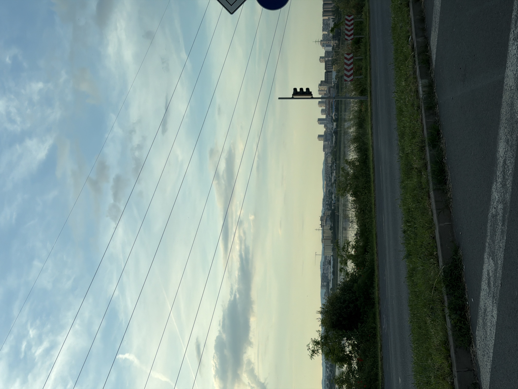 The image presents a street scene near what appears to be a river or body of water. The view includes a road with a grassy verge on one side and a pedestrian or sidewalk area on the other. A line of buildings, possibly residential or commercial, is visible in the distance across the water. Overhead power lines stretch across the sky, which is filled with clouds. There is a traffic signal or light post beside the road. Red and white chevron road signs are also present.
Without additional information, it's impossible to confirm if the image is taken in Burgas, Bulgaria.