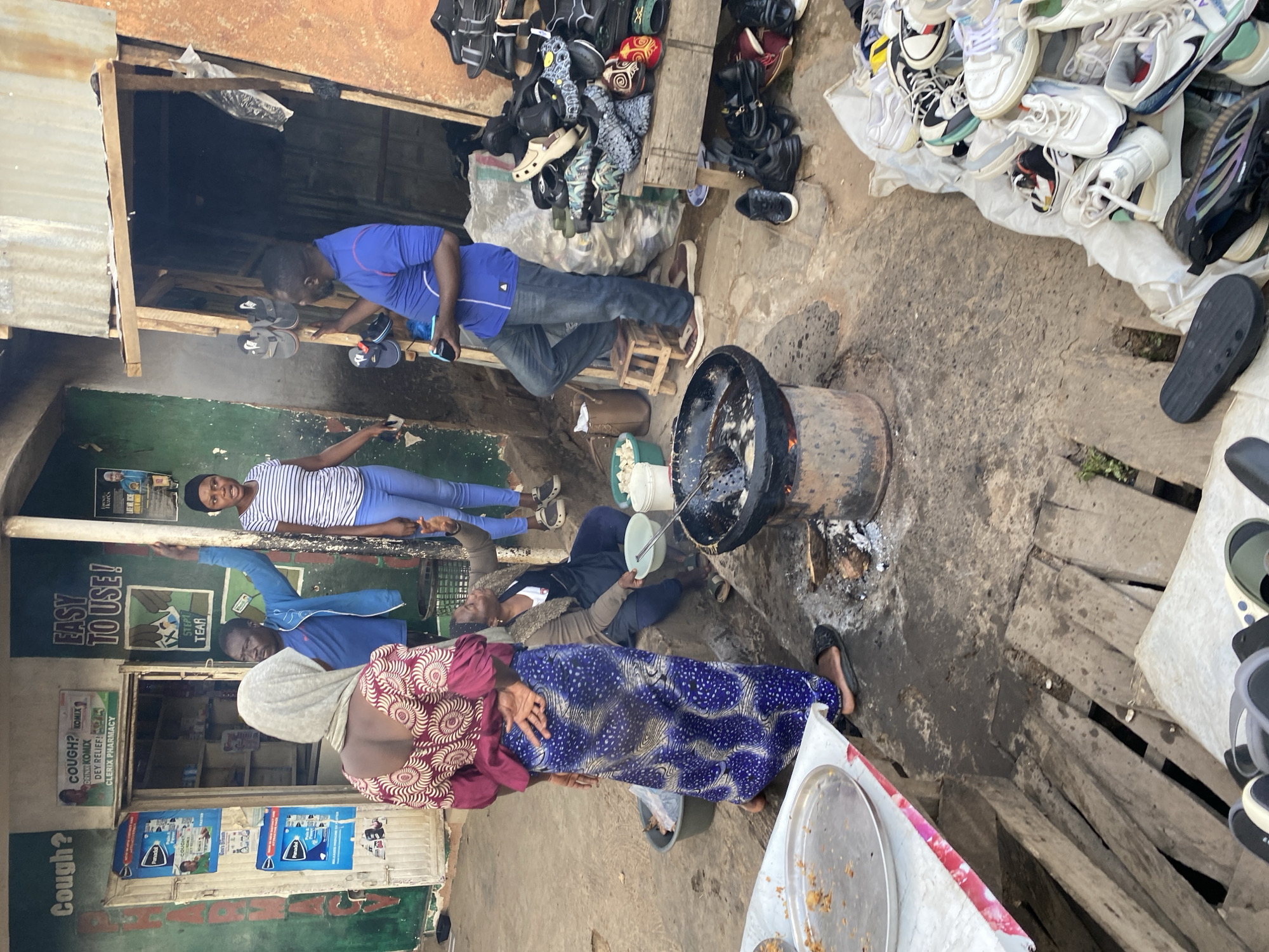 In Bukuru, Nigeria, an outdoor market scene unfolds with multiple individuals engaged in various activities. To the left, a green-painted building with signage indicating "Cough?" and "EASY TO USE!" suggests a commercial establishment, possibly a pharmacy given the visible product advertisements on the wall.
In the foreground, a woman wearing a headscarf and a colorful patterned dress stands near a table with a large metal platter of food. Beside her, another woman in a blue headscarf and a dark apron is seated, holding a bowl. Dominating the center of the image is a large metal pot or drum on a fire pit, with cooking utensils in use, indicating food preparation. A man in a blue shirt and jeans stands near this cooking area, holding a dark object, possibly a phone, and looking towards the cooking.
To the right, a man in a blue shirt and jeans is examining footwear displayed on a wooden shelf and a pile on the ground. Various types of shoes, including sandals and sneakers, are visible. The environment appears to be a busy, somewhat cluttered outdoor area with a concrete ground surface and wooden structures. The overall impression is one of a bustling marketplace where food is being prepared and goods are being sold.