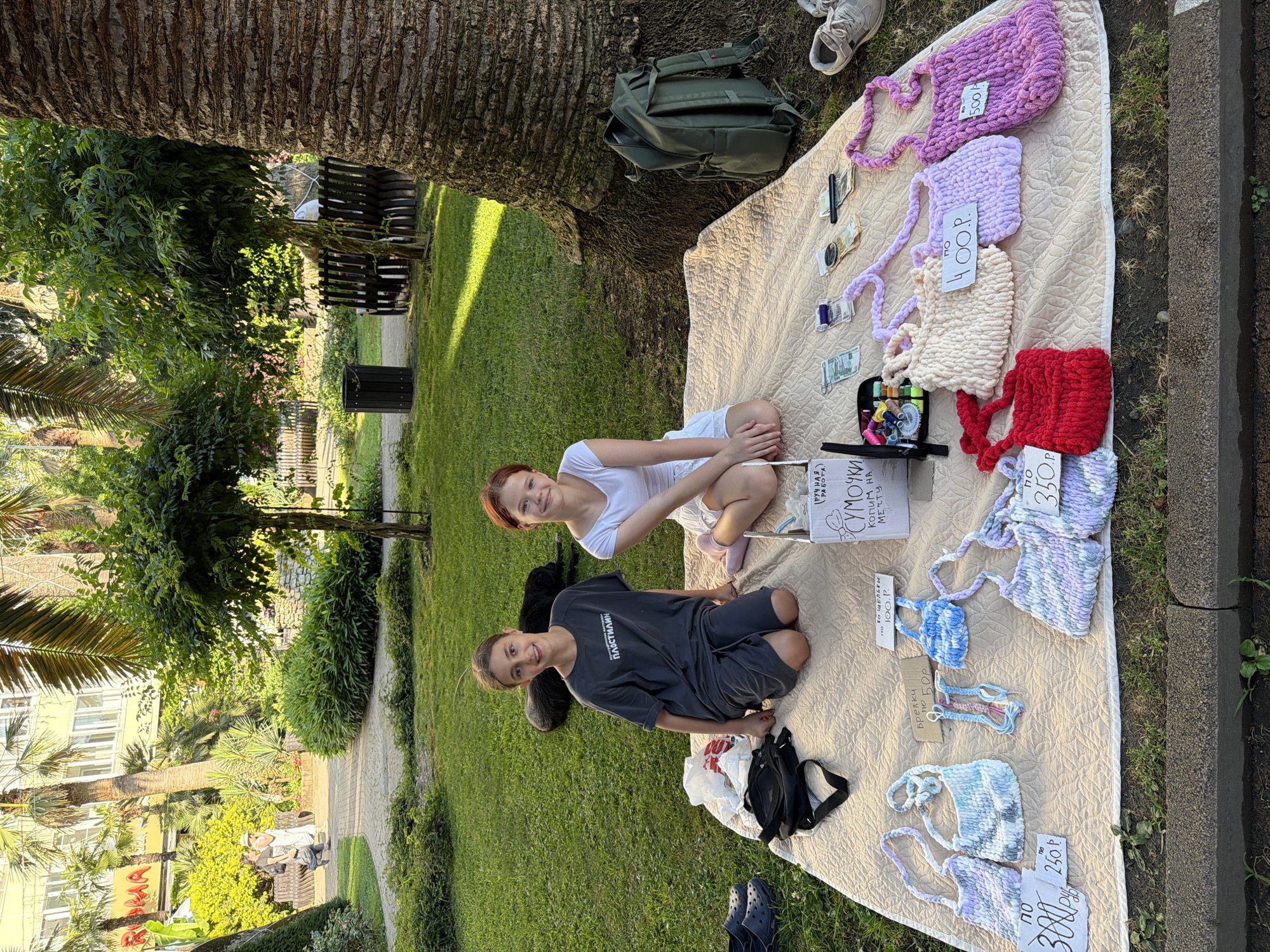 Two young people, a girl with red hair wearing a white t-shirt and shorts, and a boy in a dark t-shirt and shorts, are sitting on a blanket spread on the grass under a large palm tree. They appear to be selling handmade knitted items, including bags and keychains, displayed on the blanket. Several items have handwritten price tags with prices indicated in rubles (e.g., 500, 400, 100, 350, 250). A sign reads "СУМОЧКИ РУЧНАЯ РАБОТА КОПИМ НА МЕЧТУ" which translates to "Handmade bags. We are saving for a dream." A backpack and a sewing kit with colorful threads and needles are also visible. In the background, a pathway winds through a park with lush green grass, more palm trees, and buildings. The lighting suggests it is a sunny day, likely daytime. The overall scene is casual and outdoors, possibly a market or a small roadside sale.
