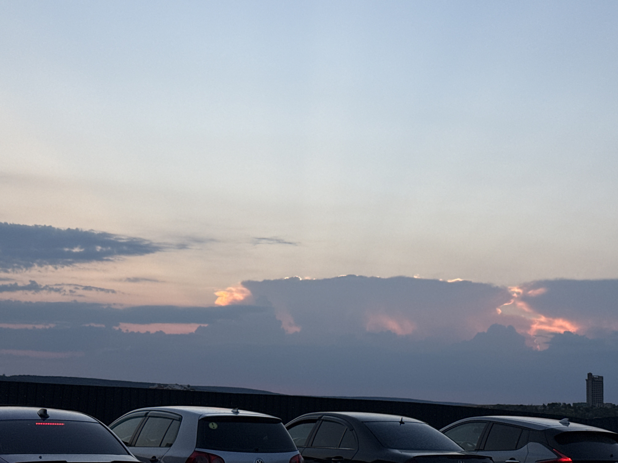 The image depicts a twilight scene with a dramatic sky filled with clouds illuminated by the setting sun, casting hues of pink and orange. In the foreground, several cars are parked in a line, their rear ends visible. The cars appear to be a mix of makes and models, including a silver hatchback and a dark sedan. Behind the cars, a dark, horizontal structure, possibly a wall or fence, runs across the frame. In the far background, a tall, slender building, resembling a high-rise or tower, stands against the horizon. The overall impression is that of a quiet evening on the outskirts of a city, possibly a parking area or roadside observation point. The specific location in Ialoveni, Moldova, is not visually discernible from the image itself, but it is described as a scene likely captured from a vantage point overlooking a landscape on the edge of the city.