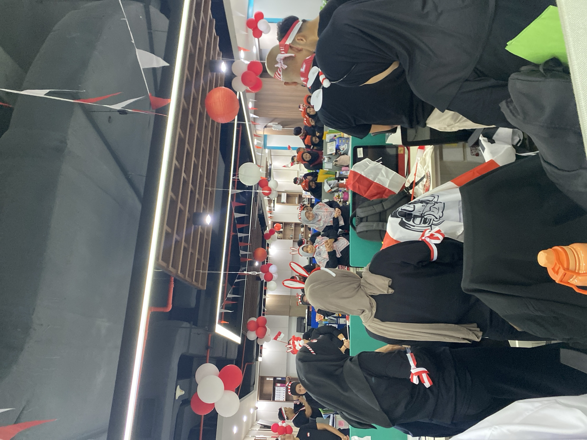 The image depicts an indoor gathering of people, likely colleagues, in an office environment. The scene is decorated with red and white motifs, suggesting a celebration or national event. People are wearing red and white headbands, armbands, and some have bunny ears. Balloons in red and white are suspended from the ceiling, and small red and white flags are strung across the room. In the foreground, several individuals are visible from behind, some wearing black attire and headscarves. In the background, more people are gathered around desks and cubicles, some appear to be standing or interacting. The overall atmosphere seems festive and communal. The image is taken from a slightly elevated perspective, looking down into the space. The location appears to be an office space in Central Jakarta, Indonesia, given the prevalence of red and white decorations, which are the colors of the Indonesian flag, often used to celebrate national holidays.