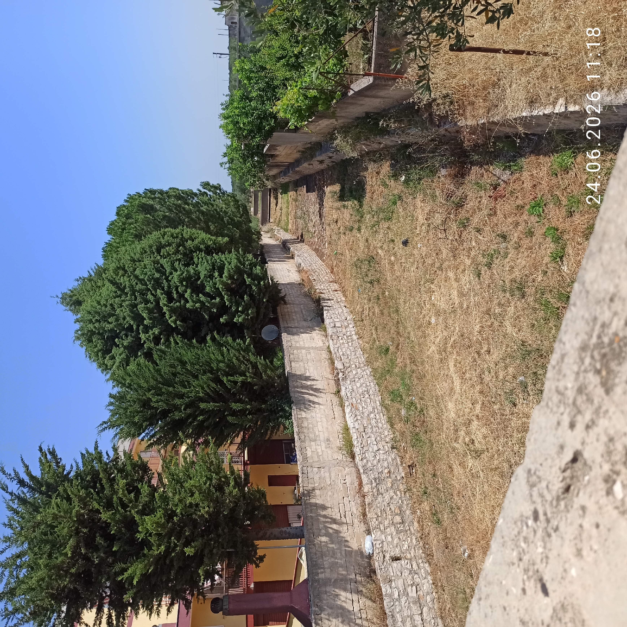 The image shows a pathway lined with stone walls and vegetation, likely in a park or a residential area. The pathway is made of brick and is somewhat uneven, with steps visible in some sections. To the left of the pathway, there are large evergreen trees, casting shadows on the ground. On the right side, the terrain is dry and covered with grass and some scattered greenery, bordered by a stone wall. In the background, buildings with yellow and red facades are visible, along with more trees. The sky is clear and blue, suggesting a sunny day. The presence of buildings and trees indicates an outdoor, possibly urban or suburban, setting. There are no people or obvious activities taking place in the image. The time of day appears to be late afternoon, given the angle of the shadows. A timestamp "24.06.2026 11:18" is visible in the upper right corner.