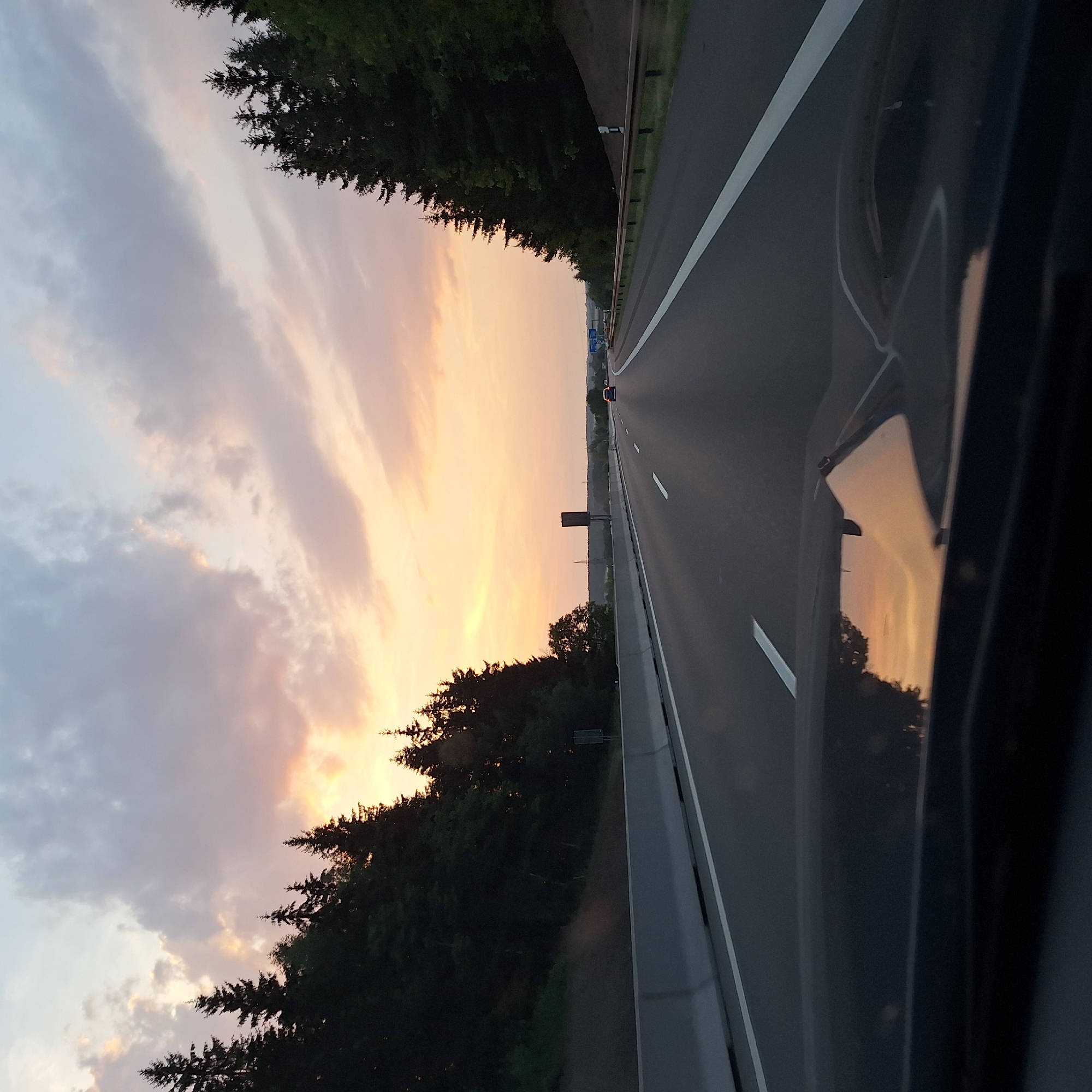 This is a forward-facing, medium shot taken from a vehicle on a highway at dusk. The sky is filled with soft, wispy clouds tinged with orange and yellow from the setting sun. On either side of the highway, dense evergreen trees line the slopes, silhouetted against the sky. The highway itself is dark asphalt, marked with white lines indicating lanes. In the foreground, the reflective surface of the vehicle's hood shows a distorted reflection of the sky and trees. A gray concrete barrier separates the highway lanes from the embankment on the left. A few cars are visible on the road ahead, moving in the same direction. The overall impression is one of a serene drive through a wooded landscape as day turns into night.