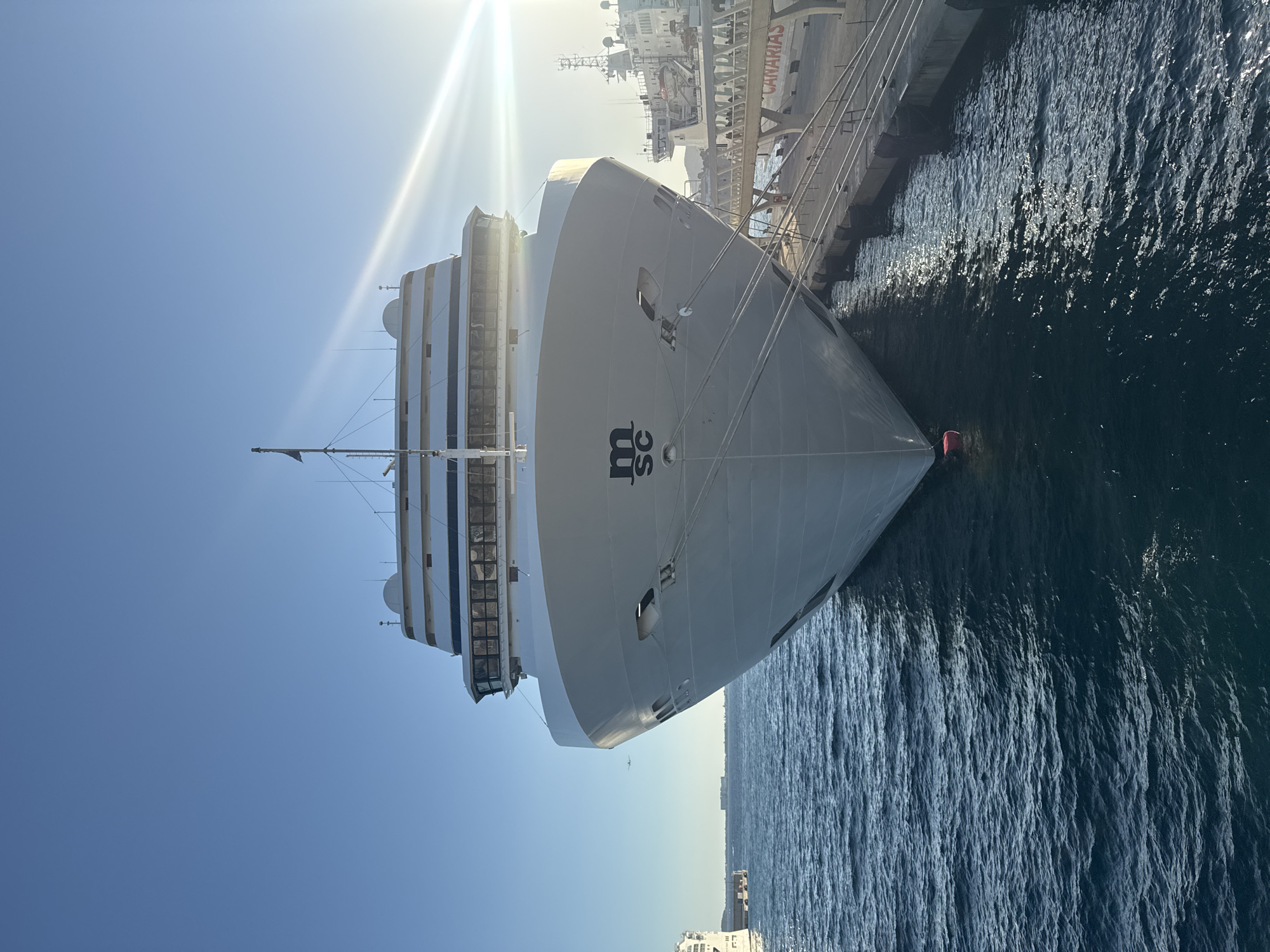 A front-facing, low-angle, medium shot captures the bow of a large white cruise ship docked at a port under a clear, bright blue sky. The sun glints off the water and the upper decks of the ship, creating lens flares. The cruise ship, identified by the "MSC" logo on its hull, is secured to a concrete pier with thick ropes. The pier has bollards and is lined with people walking and observing. In the background, another ship and port infrastructure are visible, including a sign that reads "CANARIAS." The water is a deep blue with gentle ripples. The overall scene depicts a sunny day at a busy port.