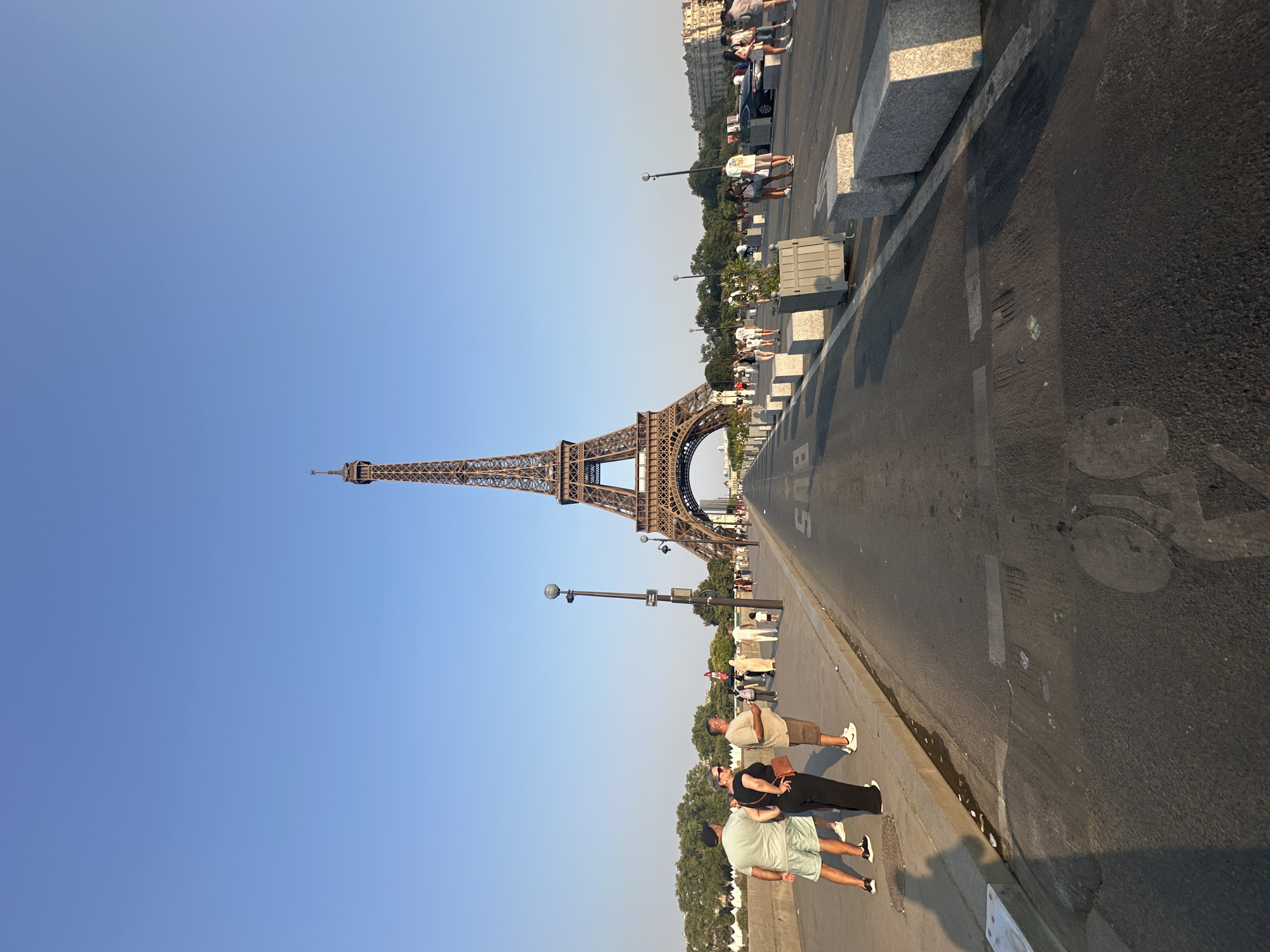 The image captures a perspective shot looking towards the Eiffel Tower in Paris, France, during the day under a clear blue sky. The Eiffel Tower is centrally located, dominating the background. In the foreground, a wide asphalt road, possibly a bridge or a street, stretches towards the tower. The road features markings for vehicles and a bicycle lane symbol.
On the left side of the road, a sidewalk is populated by several individuals. Three people, two men and a woman, are standing and looking away from the camera, engaged in conversation. Further down the sidewalk, more people are walking, some appearing to be tourists.
On the right side of the road, concrete planters and large concrete blocks are arranged as barriers. More pedestrians are visible walking along this side. A black car is parked or driving on the right side of the road.
The environment suggests a public space, likely a popular tourist area, with a mix of locals and visitors. The lighting indicates it is either morning or late afternoon, with long shadows cast by some of the people on the sidewalk, particularly visible in the lower left corner of the image. The overall scene is one of a bustling urban environment with a significant landmark as its centerpiece.