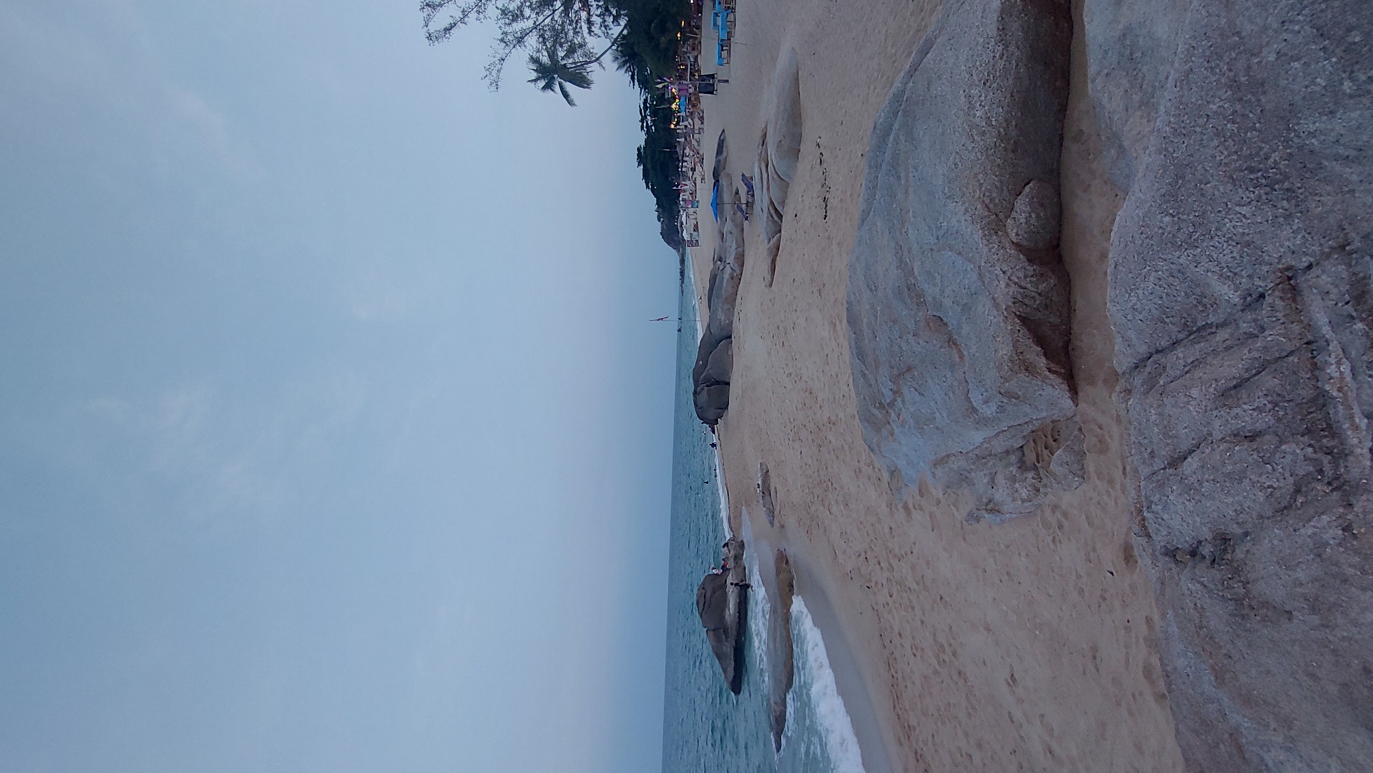 This image captures a wide shot of a tranquil beach scene, likely taken during late afternoon or dusk, given the soft, muted lighting and the subtle glow of lights along the distant coastline. The sky, a uniform light blue-grey with minimal cloud cover, dominates the upper left portion of the frame.
The central part of the image features a vast expanse of calm, blue-grey ocean water, meeting a sandy beach on the right. Gentle waves lap at the shore, creating a thin line of white foam. Large, light-colored rocks are scattered along the sandy beach, prominently featuring in the foreground and midground on the right side of the image, their textured surfaces contrasting with the smooth sand.
Along the distant coastline, a strip of dense green trees, including visible palm trees, lines the beach. Behind the trees, a series of structures, possibly resorts or restaurants, are visible, marked by small, colorful string lights, suggesting evening activity. Scattered along the distant beach and in the shallow water are several small figures, indicating people are enjoying the beach, some appearing to relax on beach chairs or stroll along the shore. A tall, slender pole with a flag-like banner is visible in the water further down the beach.
The overall atmosphere is peaceful and serene, typical of a relaxing coastal setting. There is no visible text in the image.