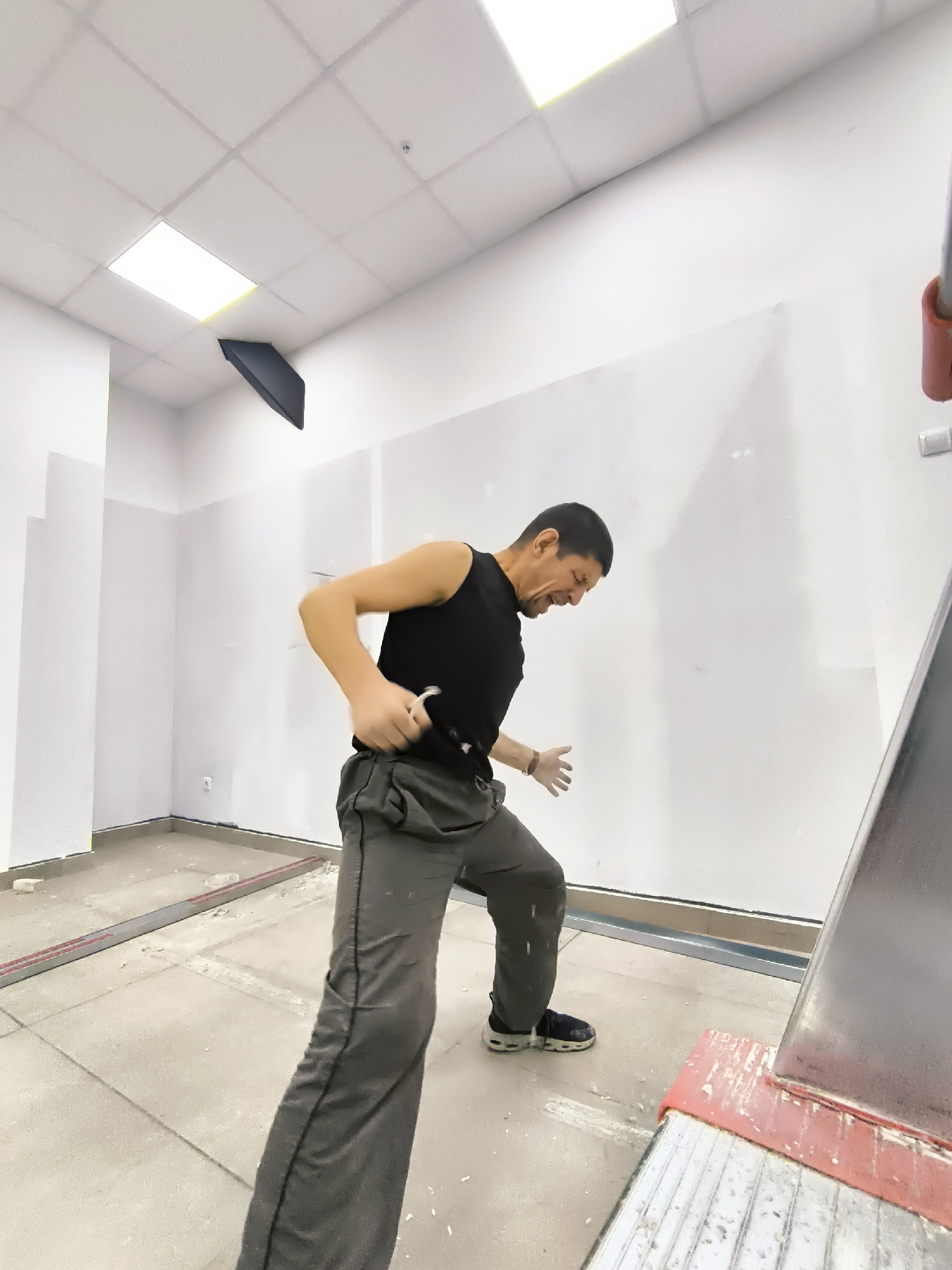 A man in work clothes, a black tank top and gray pants, is actively engaged in what appears to be construction or renovation work. He is positioned in the center of the frame, mid-motion, with his right arm raised as if to strike something with a tool held in his hand, which is partially obscured. His facial expression is strained, suggesting exertion or frustration. His left hand is extended forward, palm open.
The setting is an indoor space, likely a room under construction or renovation, characterized by bare walls, a tiled or concrete floor with debris, and a suspended ceiling grid. A large, rectangular light fixture is illuminated on the ceiling, and a dark, angled object hangs precariously from the ceiling in the upper left quadrant of the image. A metallic structure, possibly part of scaffolding or equipment, with red accents, is visible on the right side of the frame. The walls are predominantly white or very light gray, with a dark border along the base.
The scene appears to be during the daytime, inferred from the bright lighting in the room. There are no visible weather cues, as it is an indoor environment. The overall impression is one of a labor-intensive task. The man's posture and expression convey a sense of intense effort. The location cues within the image are minimal, but the context provided places the scene in the Qarağandı city administration building in Kazakhstan. No visible text is present in the image.