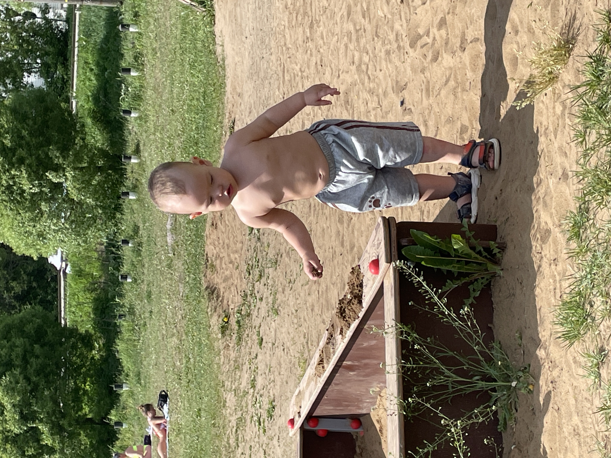A young child stands on a sandy surface next to a wooden structure with red spheres. The child is shirtless, wearing gray shorts and sandals. The structure appears to be a sandbox or play element, with greenery growing nearby. In the background, there is a grassy area with trees and other people visible in the distance. The location is Perm, Russia.