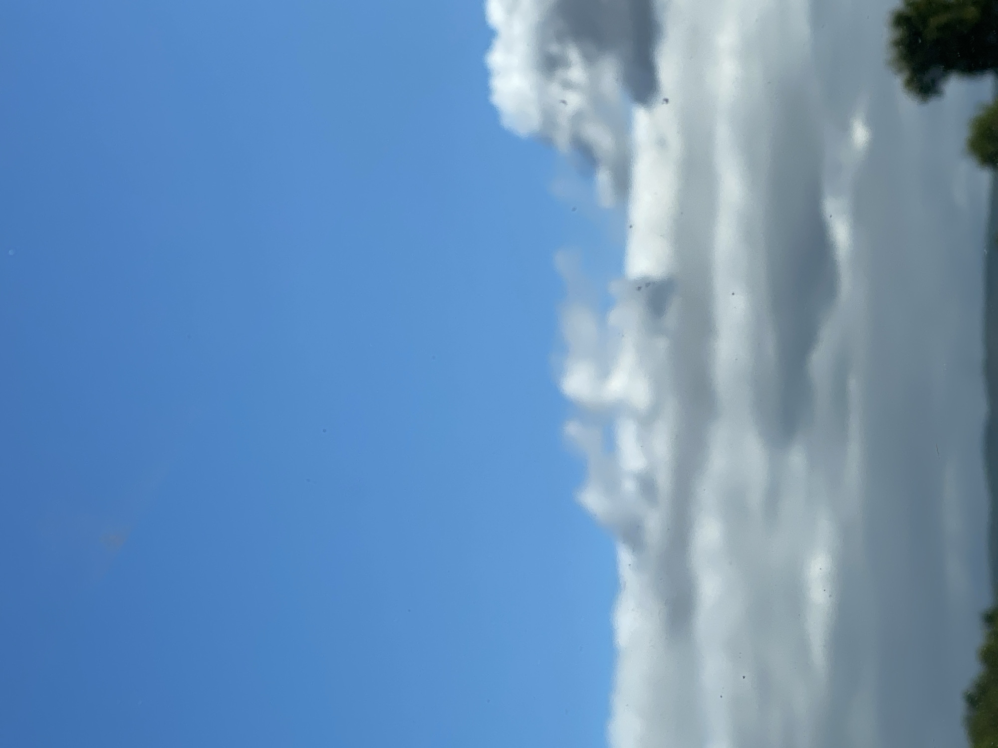 A view of the sky over Nordhausen, Germany, shows a clear blue sky with scattered clouds. The clouds are white and gray, with some appearing fluffy and others more wispy. The bottom of the image shows a blurred green landscape, possibly trees and hills. The image is taken from a slightly elevated perspective, looking up at the sky.