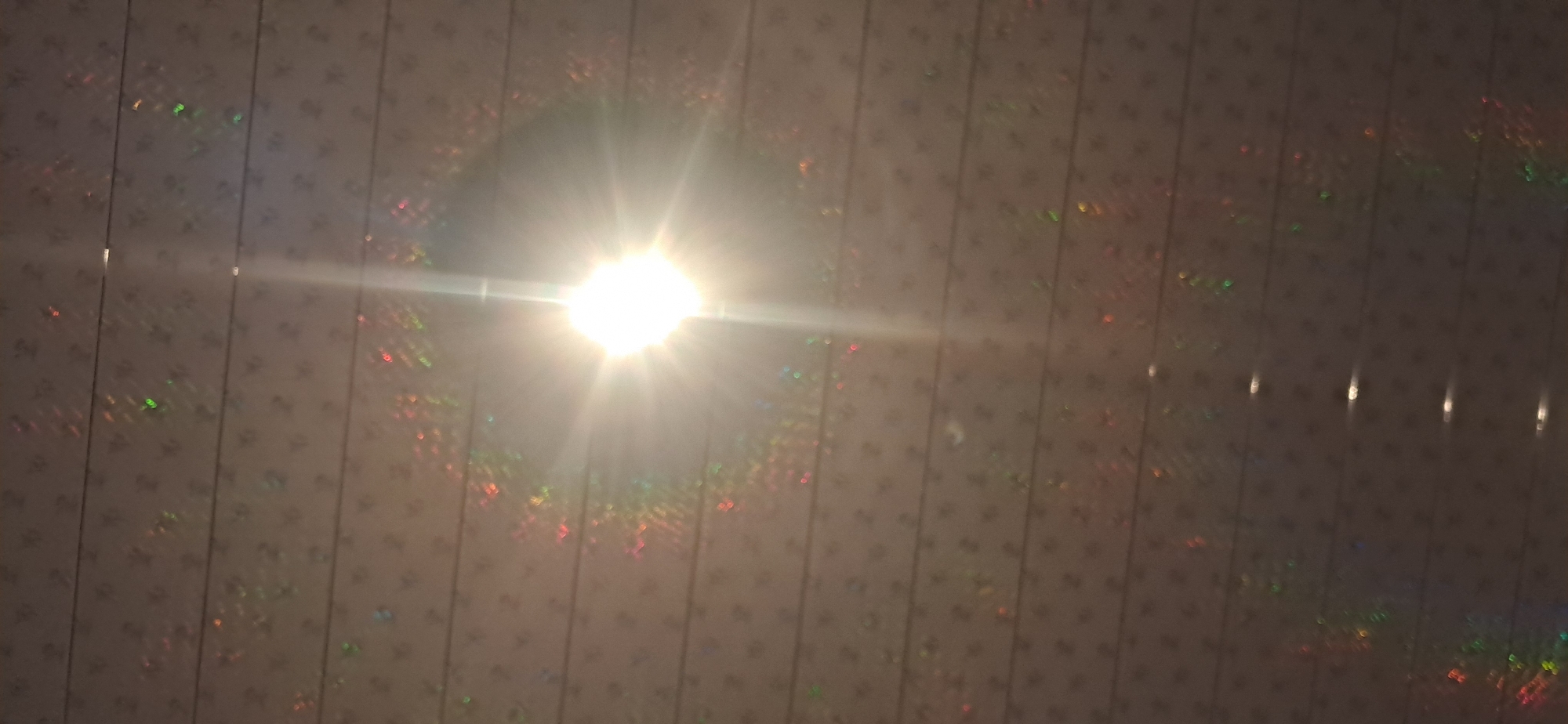 The image depicts a bright light source, likely the sun, with visible light flares and a halo effect, illuminating a textured, patterned surface. The surface appears to be a ceiling with horizontal lines and a repeating small, dark pattern, possibly flowers or stars, consistent with decorative paneling. There are iridescent reflections scattered across the surface, suggesting a holographic or glittery material. The overall lighting creates a warm, slightly hazy atmosphere. The specific location in the city of Yazy, Turkmenistan, is not discernible from the visual information provided in the image.