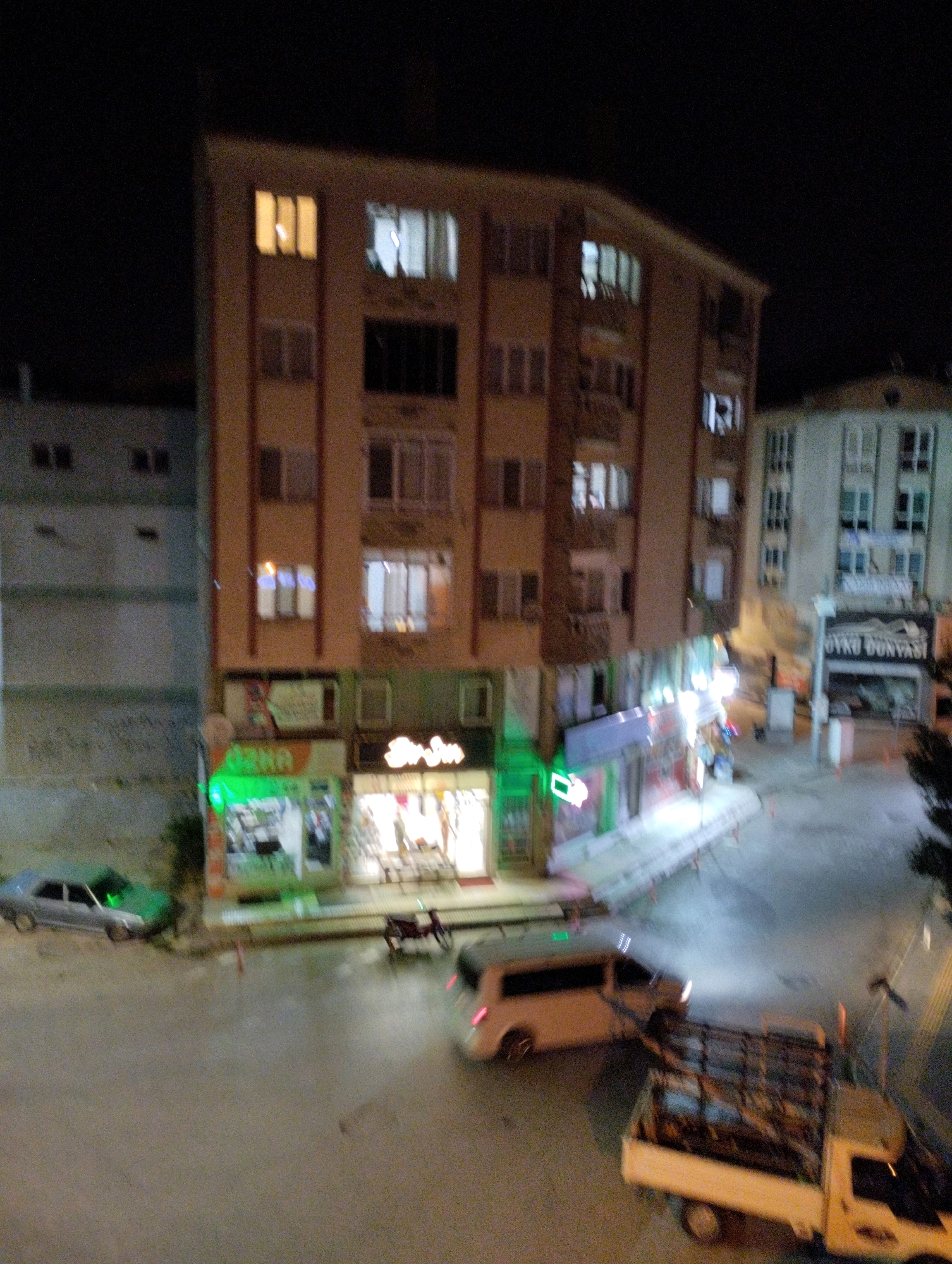 The image shows a street scene at night in Çorum, Turkey, with a focus on a multi-story building housing several businesses. The building on the right appears to be commercial, with glowing signs for "OZKA" and "Sirin" visible on the lower floors, suggesting retail establishments. Above these shops, the building has multiple levels with illuminated windows, indicating residential units. To the left of this building is a lighter colored, taller structure, also with multiple illuminated windows.
In the foreground and on the street, several vehicles are present. A light-colored van or minivan is seen driving away from the viewer, its headlights on. A truck with a caged bed is parked to the right of the van. Further to the left, a dark-colored car and a green car are parked on the street. The street itself is paved, and the overall impression is of a somewhat busy urban environment at night. The lighting from the businesses and vehicles illuminates the street, creating reflections on the wet pavement. The sky is dark, typical of nighttime. There are no visible people in the image, but the presence of lit windows and businesses suggests activity within the buildings.