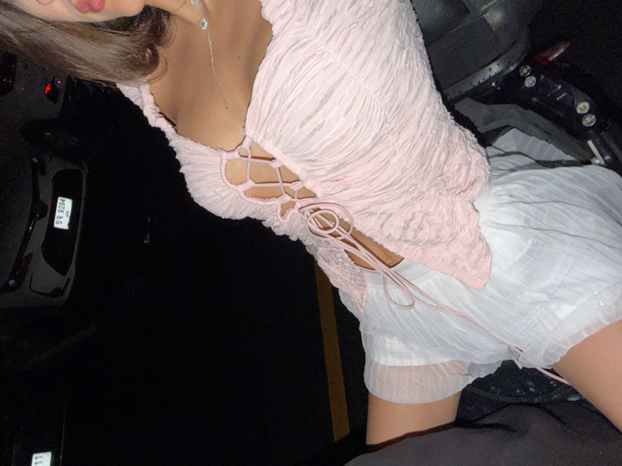 The image shows a woman in a pink textured top with a lace-up front and a white pleated skirt. She is possibly inside a car, given the presence of a car dashboard on the right and a car door on the left. The location information (Choeng Thale, Thailand) is extraneous to the content of the image.