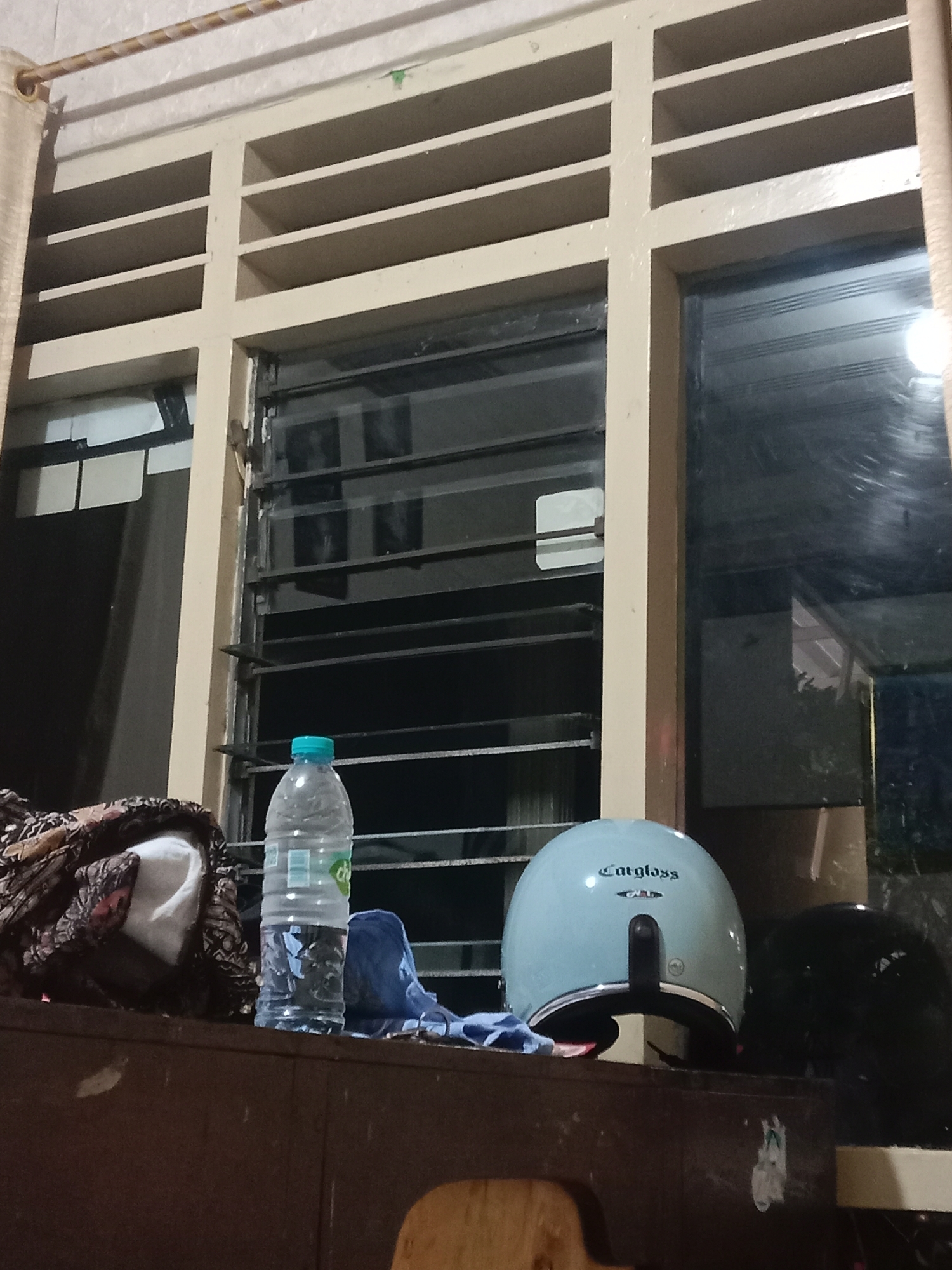 An interior scene, possibly a room in Madiun, Indonesia, is depicted at night. In the foreground, a dark brown wooden surface holds several objects: a patterned fabric item with a white cloth peeking out, a clear plastic water bottle with a blue cap, a blue piece of fabric, a light blue helmet with "Cargloss" branding, and a black helmet. Behind these items, a large window with horizontal louvers is visible. The window reflects the indoor lighting and shows the exterior environment dimly. The overall lighting is dim, suggesting it is nighttime or the room is not well-lit.