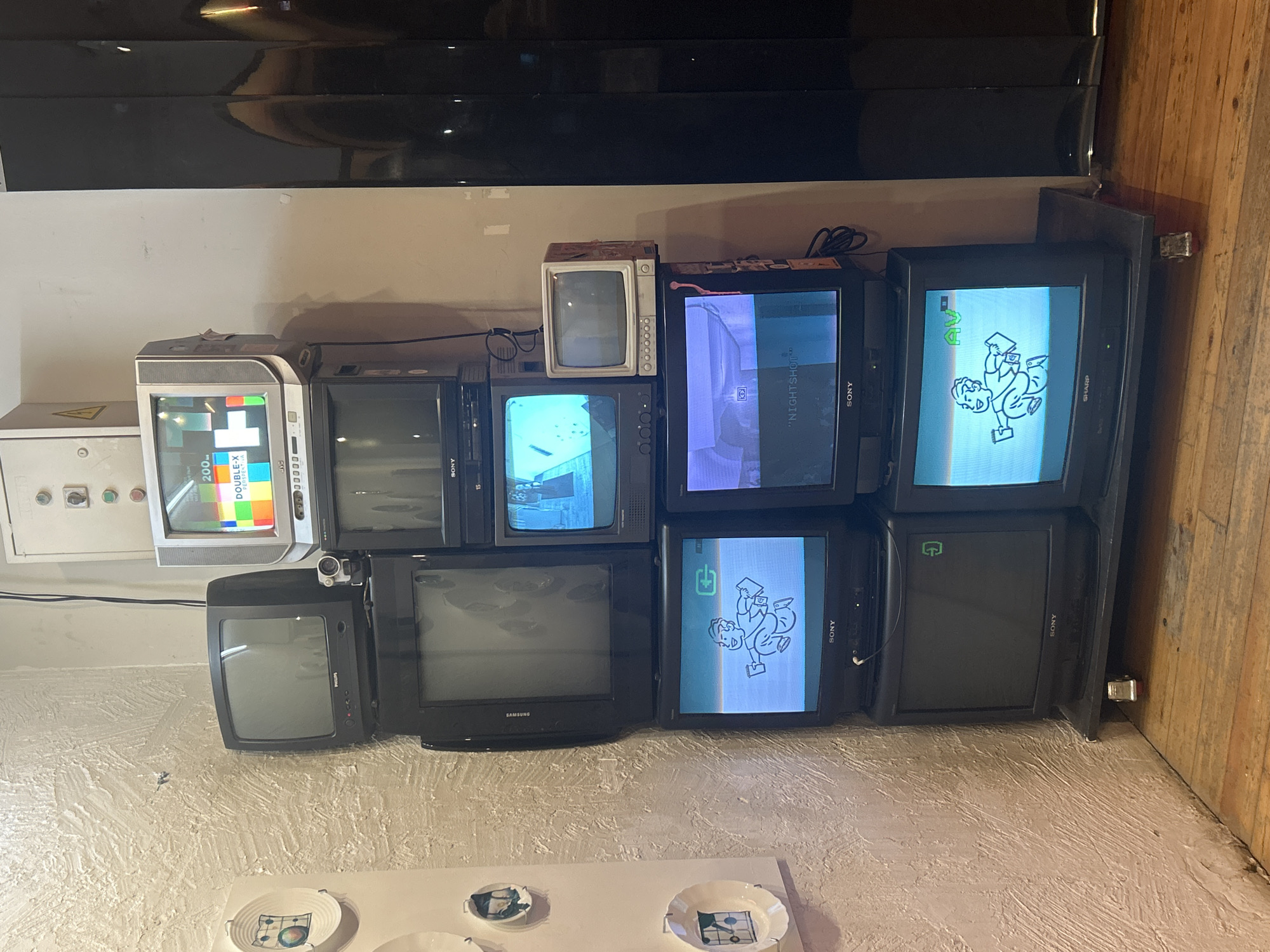 The image shows a collection of old cathode-ray tube (CRT) televisions stacked against a wall in what appears to be an indoor setting, potentially an exhibition space. At least nine TVs are visible, with some displaying static or images related to television testing, while others show a character in a cartoon style. The TVs vary in size and brand, including Sony, Sharp, Samsung, and JVC. Some of the screens display what is written in Russian. Additionally, there is a white electrical box to the left with a hazard symbol on it. In the foreground, there are some plates with images matching the images on the TV screens. There is a wooden floor to the right of the TVs. Based on the brands and languages displayed on the TV screens, it is likely in Moscow, Russia.