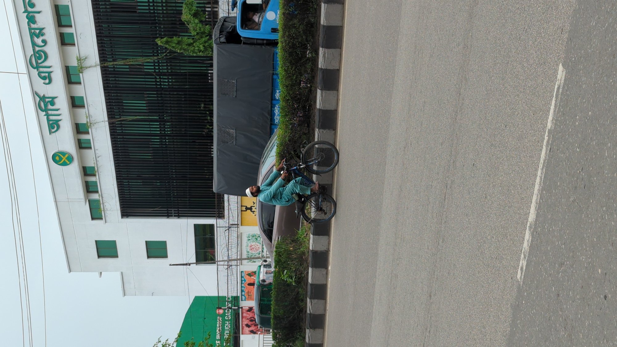 The image depicts an urban street scene, likely in Dhaka, Bangladesh, during daytime under an overcast sky. A man, wearing a light teal-colored tunic and a white prayer cap, is seated on a black bicycle near the edge of an asphalt road. He is positioned next to a black and white checkered curb, behind which a strip of green bushes separates the road from the adjacent buildings.
Further down the road, a light blue truck with a dark canvas cover is visible. White Bengali script on the truck's side reads "মেট্রোপলিটন পুলিশ" (Metropolitan Police), suggesting it is a police vehicle. A brown car is partially visible between the man and the truck.
On the left side of the frame, a multi-story white building with numerous green-tinted windows dominates the background. Prominently displayed on its facade is large green Bengali text, which reads "আঁখি মণি টেলিভিশন" (Akhi Moni Television), alongside a circular green logo featuring stylized letters. Below this building, other structures and a green signboard with partially obscured Bengali text can be seen. The road itself is gray asphalt, with a faint white line visible.