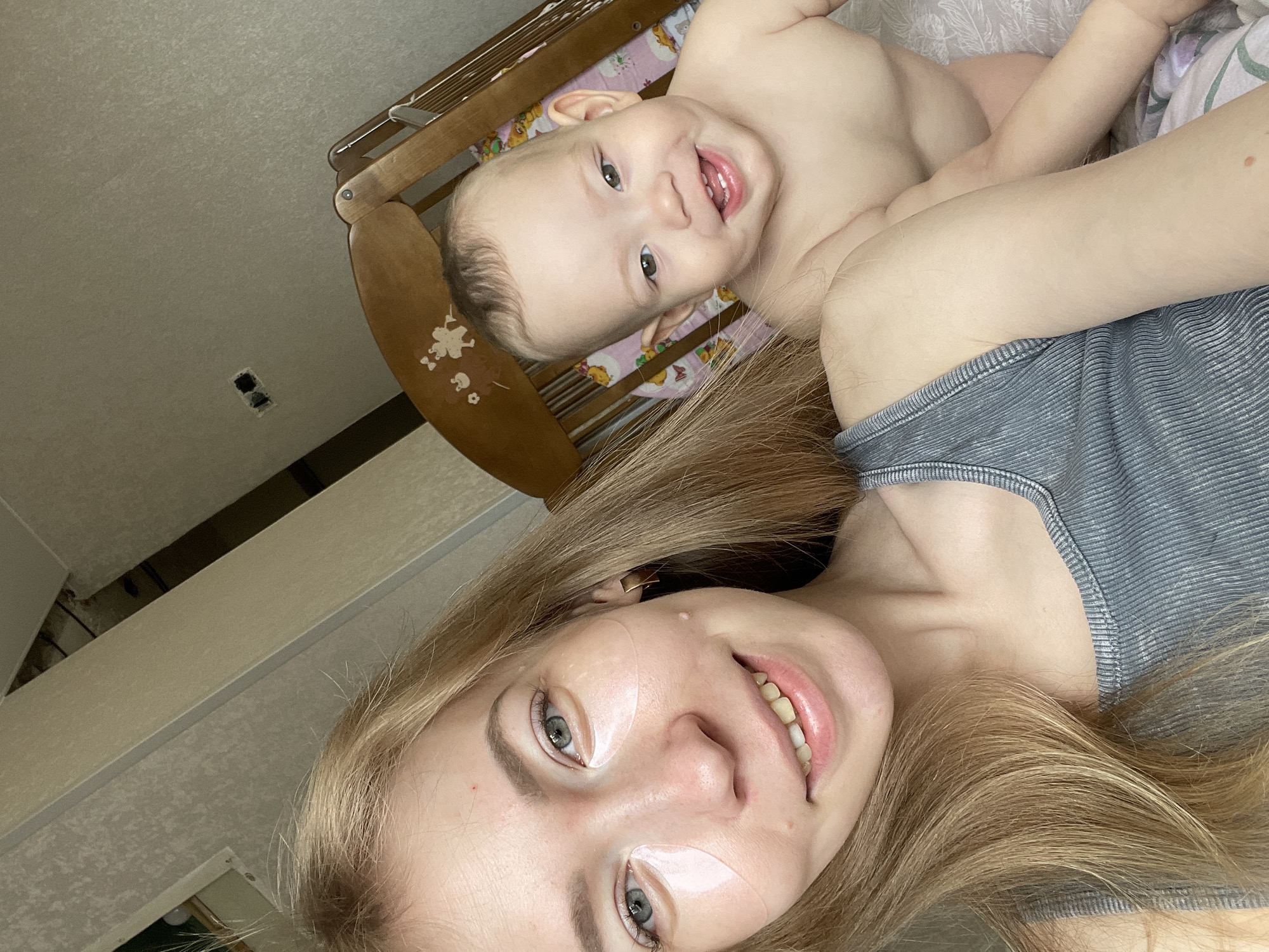 A selfie depicts a woman with long blonde hair, pale skin, and adhesive strips under her eyes. She is lying down and smiling. A baby, with light skin, is lying next to her in a wooden crib with a patterned pink and yellow mattress. The background shows a room with light wallpaper and a ceiling with exposed wiring. The location is likely a domestic setting in the city of Perm, Russia.