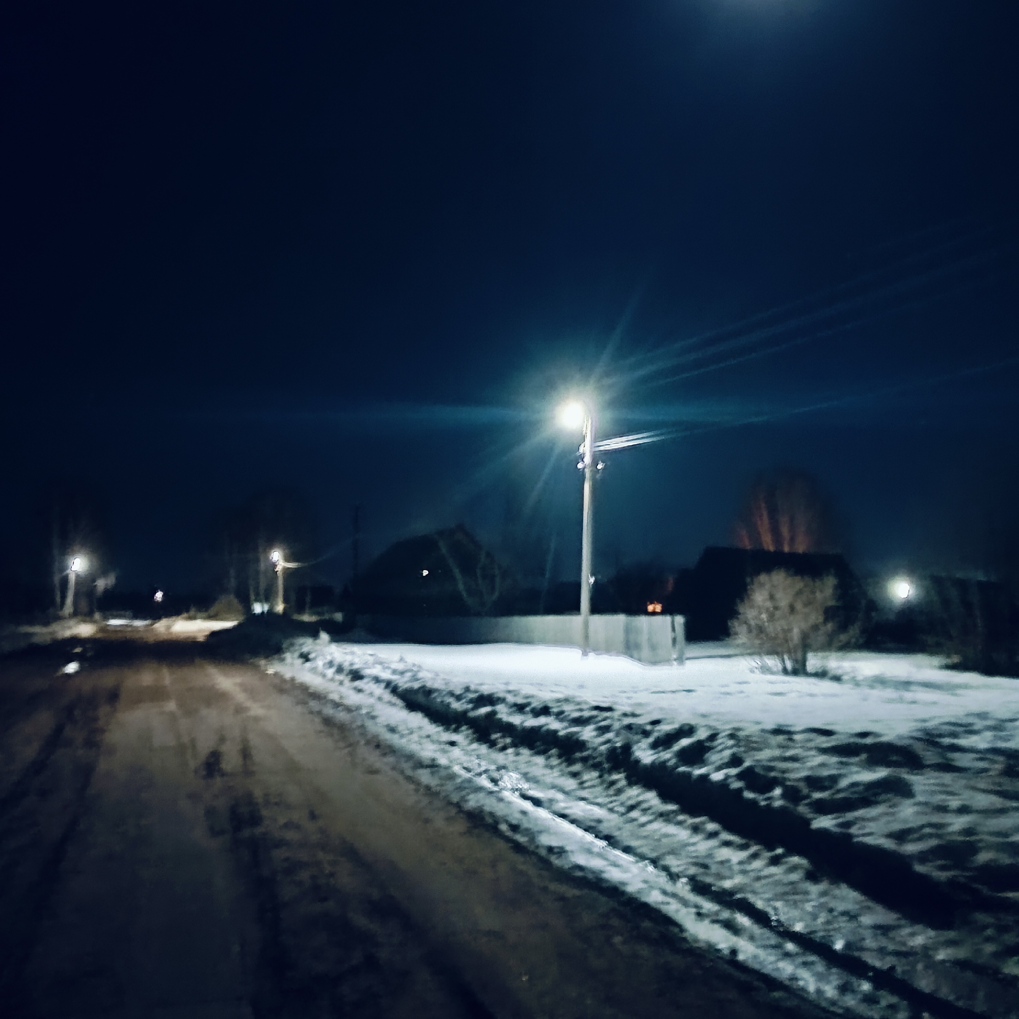 The image shows a nighttime scene on a road in Буренка, Russia. The road appears unpaved and muddy, with snow lining the sides. Streetlights illuminate the area, casting a bright glow and long shadows. Buildings or houses are visible in the background, obscured by the darkness. The overall environment suggests a rural or semi-rural setting. No people are visible in the image.