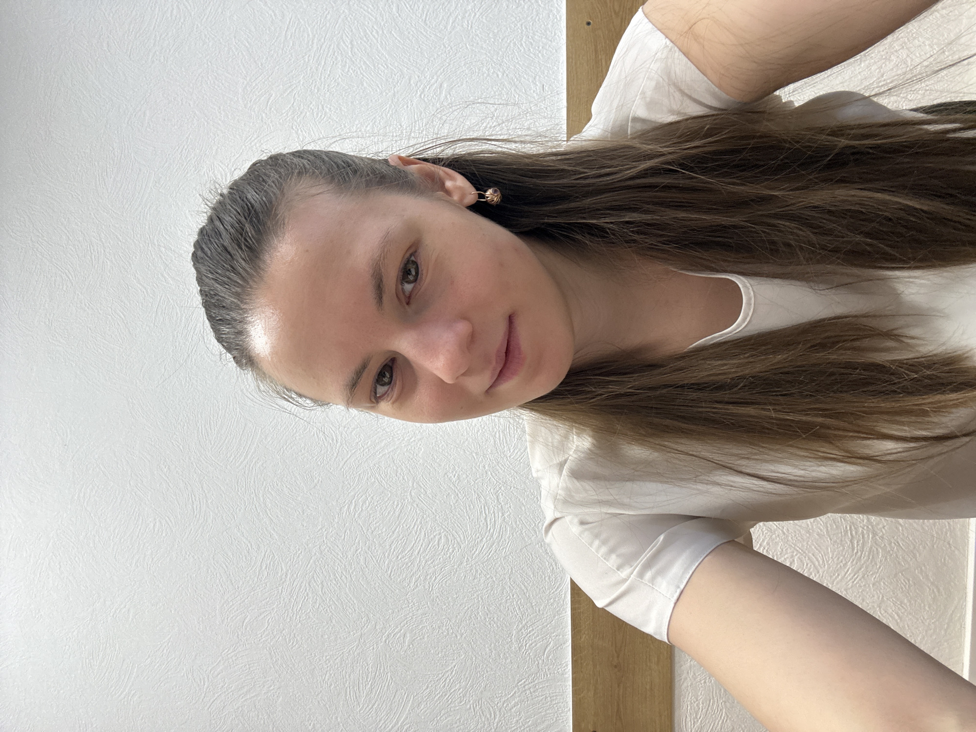 A young woman with long brown hair and fair skin is positioned at an angle facing the camera. She is wearing a white t-shirt and a small earring on her left ear. The background consists of a white textured wall and a portion of a wooden frame. The location is unspecified, and no information places the scene in Chaikovsky, Russia.