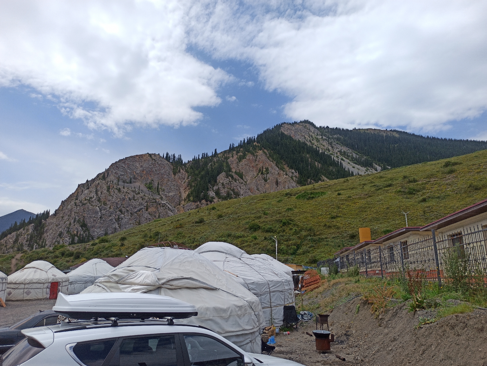 The image shows a campground with yurts and buildings in the city of Kurmeti, Kazakhstan. The scene is dominated by a large mountain in the background, covered in green grass and trees. In the foreground, there are several white yurts, some of which are covered with tarps. A white SUV with a roof box is parked in front of the yurts. There is also a building with a red roof and a black fence, and several chairs and a barbecue grill are scattered around the campsite. The sky is cloudy with some patches of blue. The overall impression is one of a remote and rustic camping location in a mountainous region.
