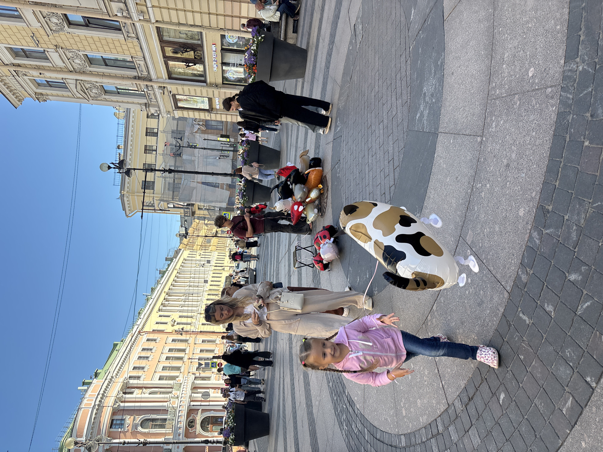 The image captures a street scene in Saint Petersburg, Russia, with the neoclassical architecture of the city visible in the background. In the foreground, a young girl with pigtails, wearing a pink hooded jacket and blue jeans, walks across a paved plaza, holding a leash attached to a large, inflatable cow-shaped balloon. To her right, a woman in light-colored loungewear stands, looking down at her phone. Behind them, a cluster of balloons, including ladybug and mushroom shapes, lie on the ground, some held by a person in a maroon shirt. Other pedestrians are visible in the midground and background, navigating the wide open space lined with potted plants and building facades. The sky is clear and blue.