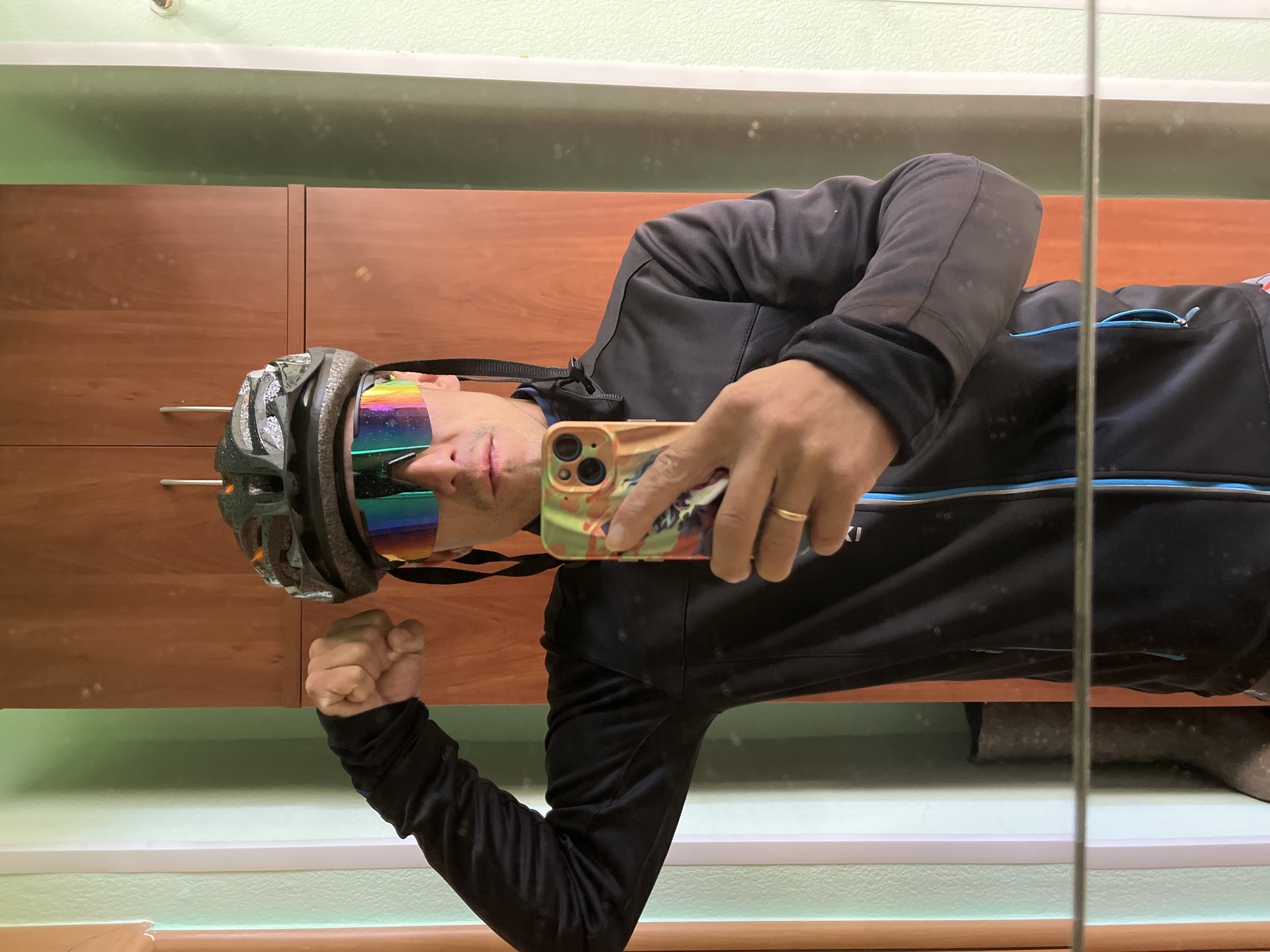An adult individual, likely male, is captured in a mirror selfie, seen from the chest up. The person is wearing a dark cycling jacket with blue accents, a dark grey cycling helmet, and rainbow-reflective cycling glasses. Their right hand holds a smartphone with a distinctive colorful, possibly abstract art-patterned case, positioned to take the photo. A simple gold-colored ring is visible on the left ring finger. The person's left hand is clenched into a fist. The background reflects a room featuring light brown wooden cabinets with silver handles and light green walls above. Part of a "KI" logo is visible on the jacket. The image captures an indoor setting, likely a home or changing area in Chaikovsky, Russia.