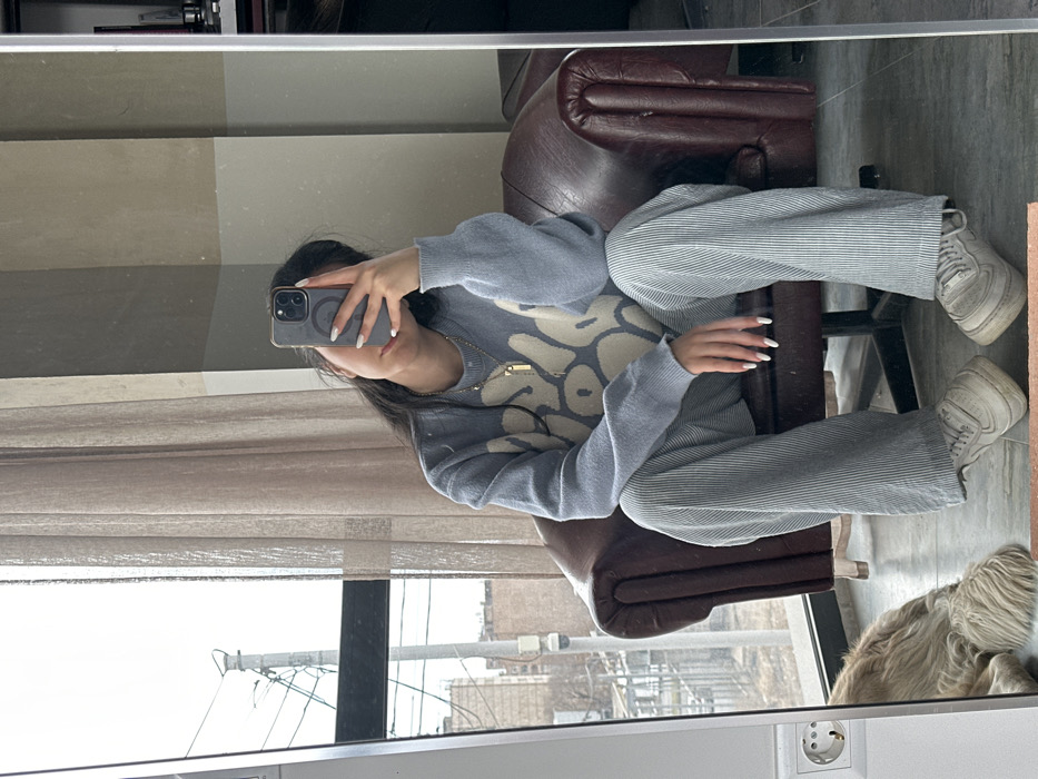 A female subject is captured in a mirror selfie, seated indoors. She wears a light blue knitted sweater with an abstract cream pattern, light gray pinstriped wide-leg pants, and off-white chunky sneakers. Her dark hair is loose, and she holds a dark gray iPhone, obscuring her face, to take the photograph. A gold-colored chain necklace with a rectangular pendant is visible. Her fingernails are manicured with white polish. She is seated in a dark brown leather armchair. To the left, a light-colored, long-haired dog is partially visible lying on the floor. The interior features large windows with light brown sheer curtains and a visible white electrical outlet on the wall. The floor is a light, possibly tiled or concrete, surface. Beyond the window, an urban landscape unfolds under an overcast sky, revealing multi-story residential buildings, utility poles with dense wiring, and an exterior-mounted surveillance camera. This scene is located in the city of Yerevan, Armenia.