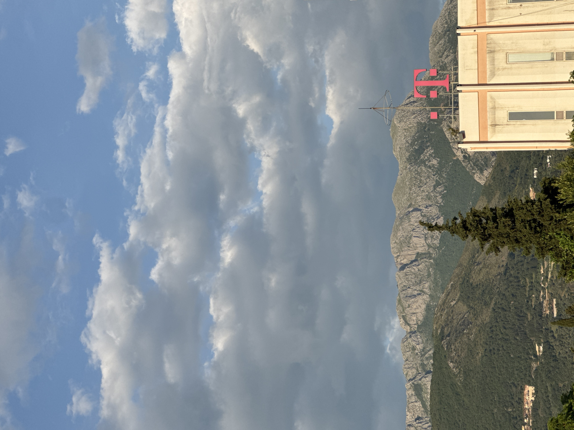 The image shows a sideways view of a cloudy sky over a mountain range in Bar, Montenegro. A large, pink "T" logo, likely for T-Mobile, is perched atop a building adjacent to the mountain. The building is beige with white trim. The foreground features green trees and foliage.