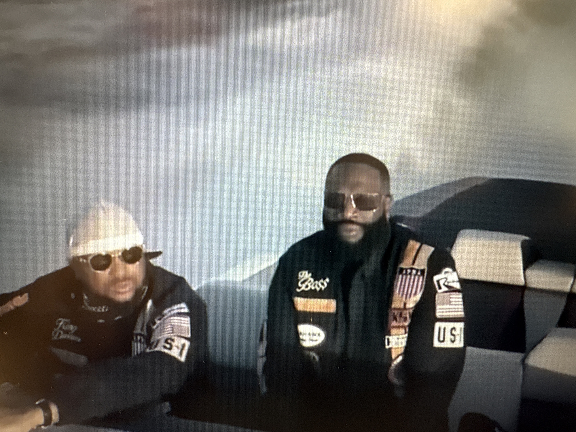In the photograph, two men are seated next to each other. The man on the left wears a light-colored cap, sunglasses, and a dark jacket with patches on the sleeve. He has his left arm resting on his lap, and his right hand is visible near a watch on his wrist. The man on the right has sunglasses and a beard, and he is wearing a dark jacket with several patches, including one that reads "The Boss." Both men appear to be in a vehicle, possibly a boat, with checkered seating visible behind them. The background is blurred and appears to be an outdoor scene with a cloudy sky. There is no information in the image to suggest the location is the city of Sofia, Bulgaria.