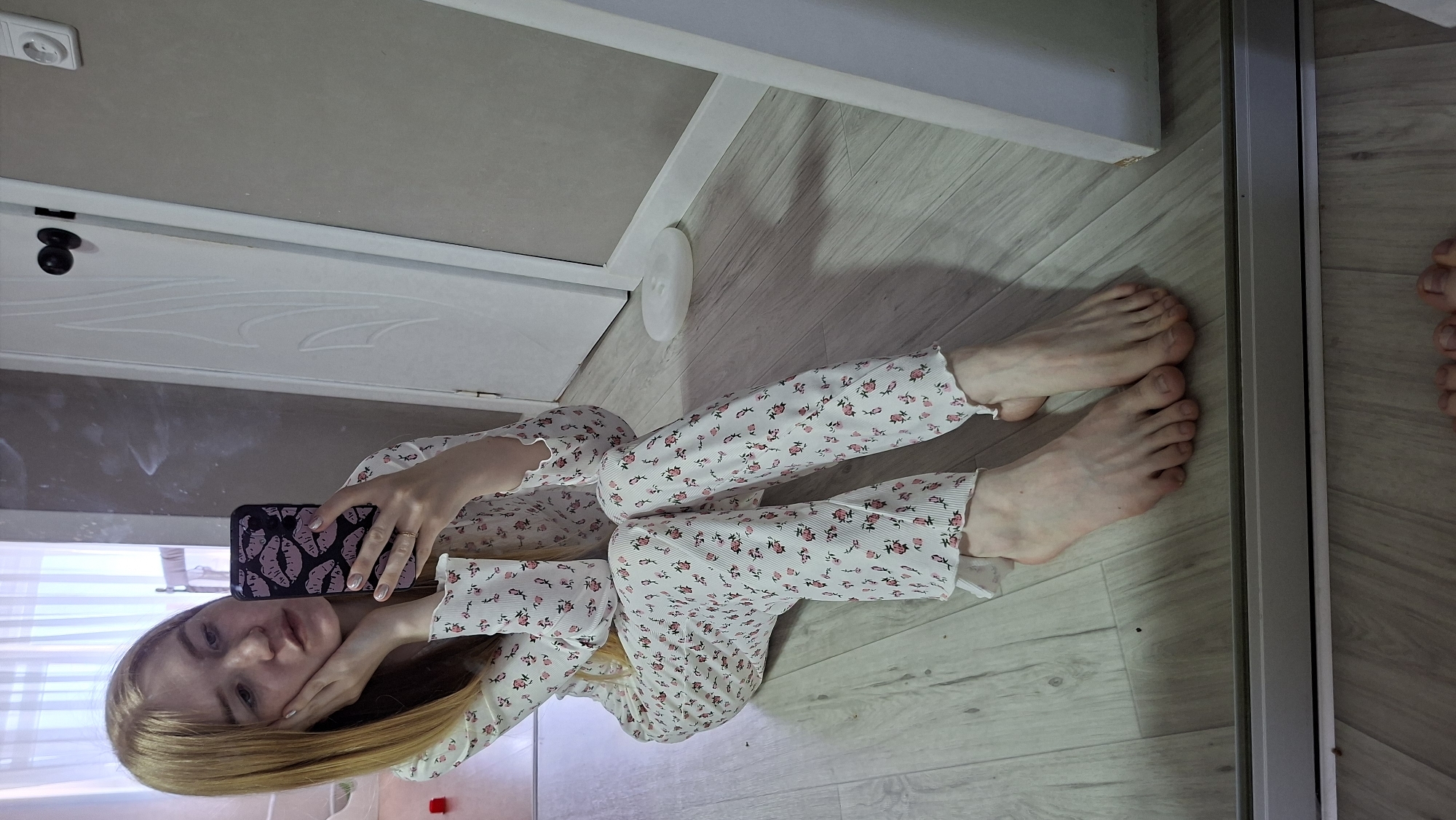A young woman with long blonde hair is lying on a light-colored, possibly laminate, floor in what appears to be an apartment in Chaikovsky, Russia. She is wearing white pajamas with a small floral print. She is holding a smartphone with a patterned case, taking a selfie. Her bare feet are visible near a mirror, which partially reflects another set of feet, suggesting a reflection. The scene includes a white door and baseboard. A white circular disc is visible near the baseboard.
