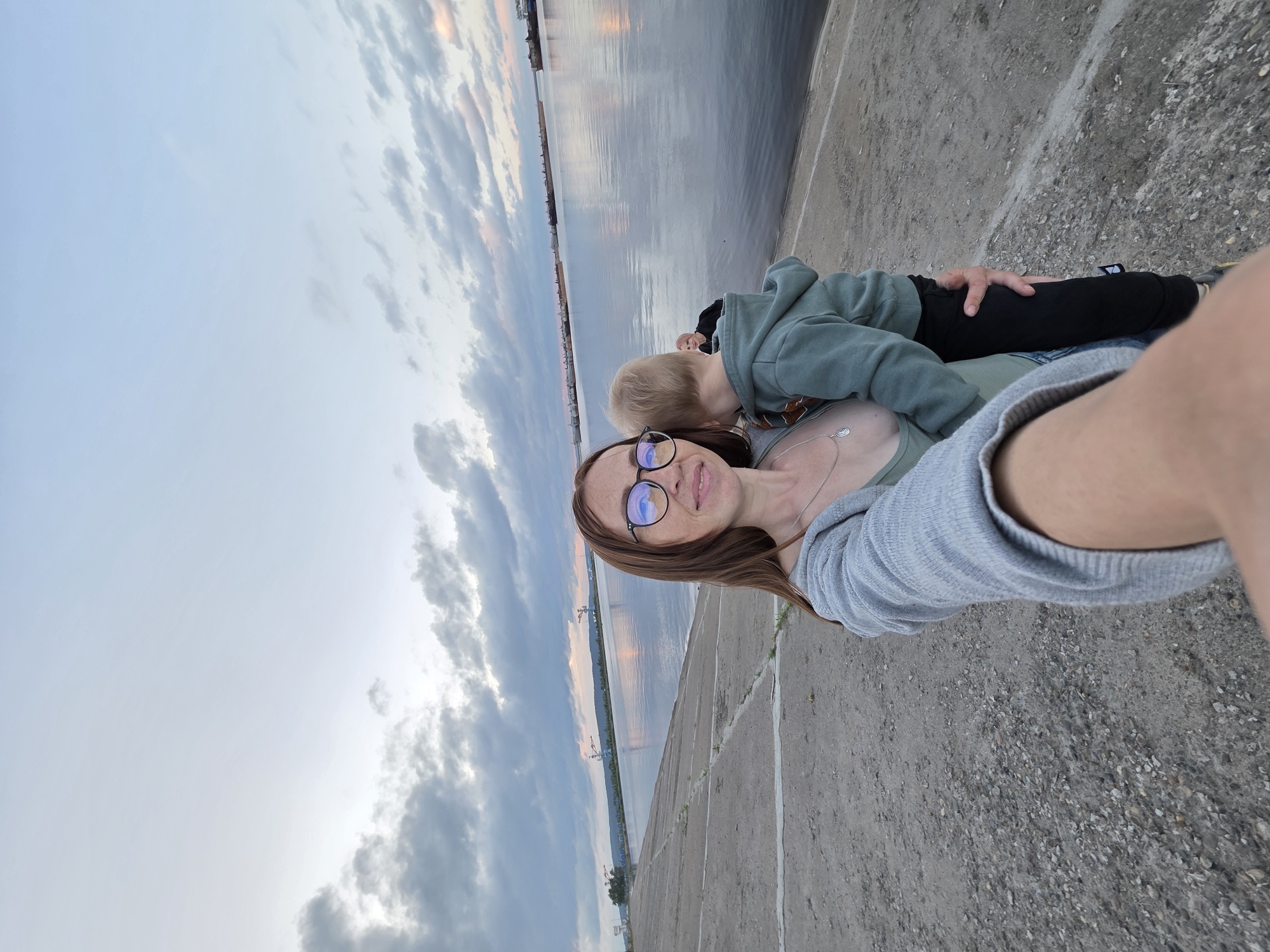 A woman is taking a selfie with a young child in her arms. The woman is wearing glasses, a grey cardigan, and a green top. The child is wearing a green hooded sweatshirt and black pants. They are standing on a concrete embankment next to a large body of water. In the background, there are several ships docked and a cloudy sky. The location appears to be the embankment of the Kama River in Chaikovsky, Russia, given the presence of industrial vessels and the general landscape typical of this area.