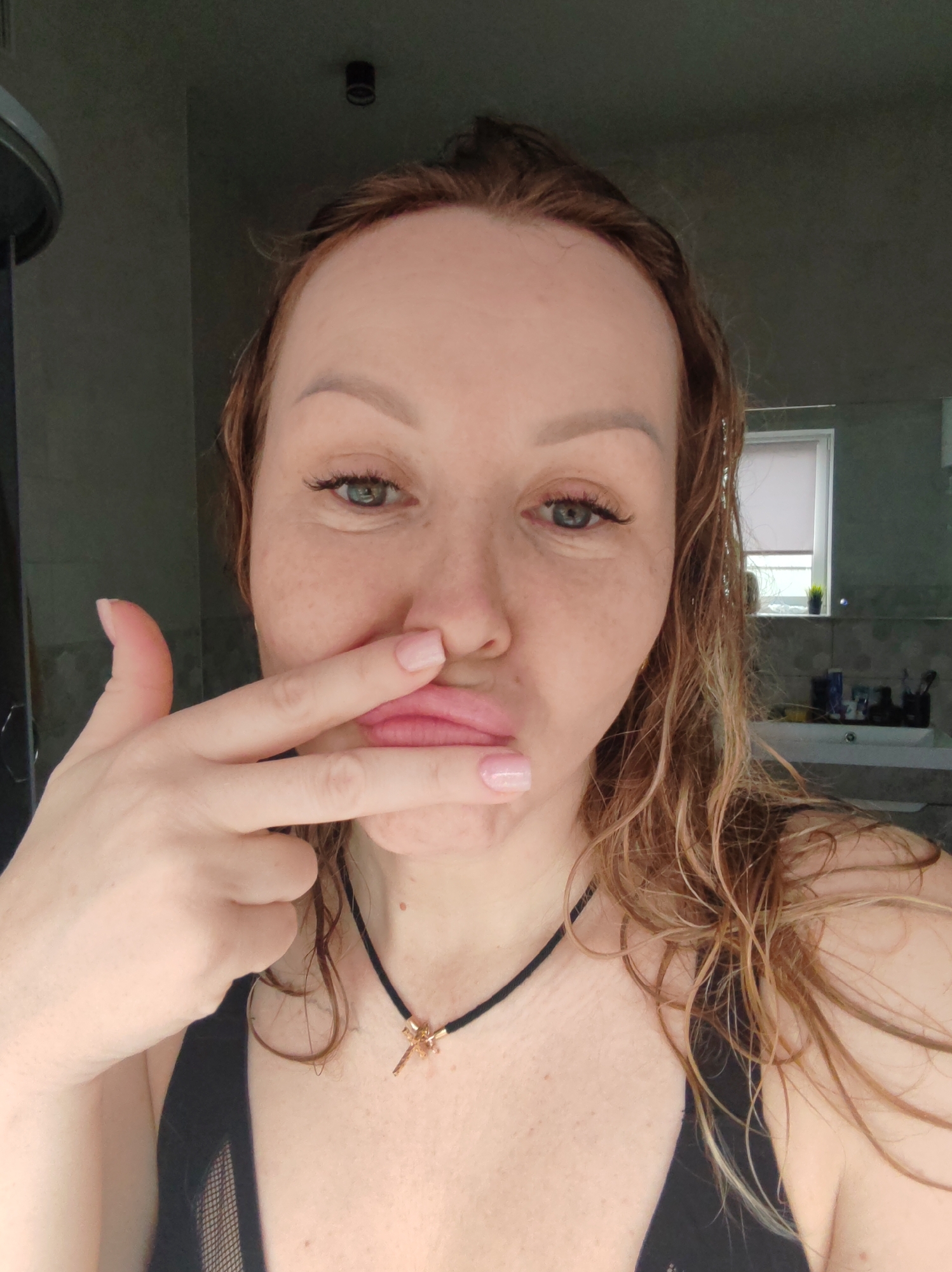 A woman is posing indoors, likely in a bathroom, based on the presence of a sink and mirror visible in the background. She has light skin, reddish-blonde wet hair, and is wearing a black top and a black choker necklace with a gold pendant. She is making a face, pressing her fingers against her mouth. There are freckles visible on her face. The background includes light grey walls, a shower stall, a mirror, and a window with a closed blind. The location is the city of Chaikovsky, Russia.