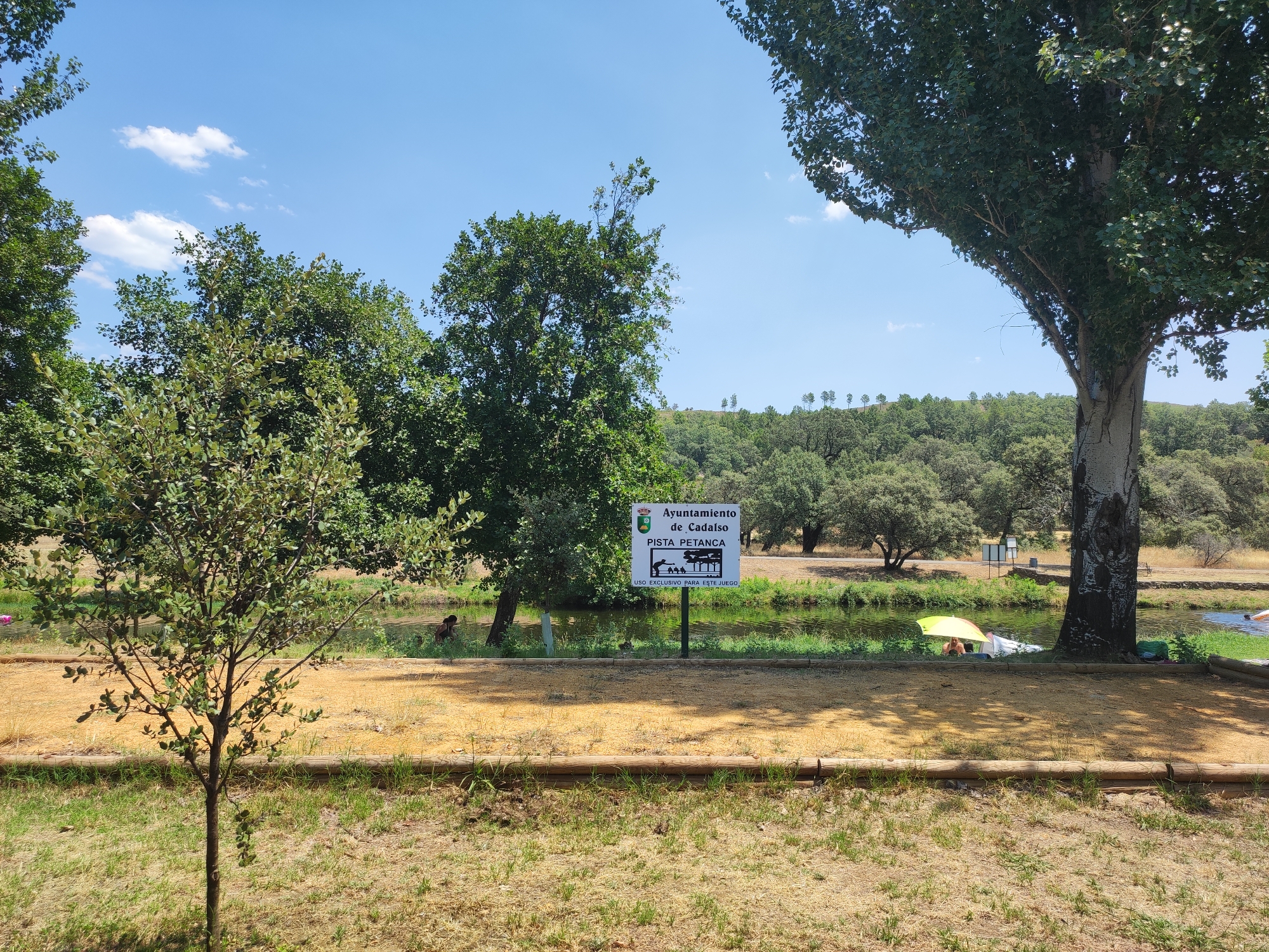 The image depicts an outdoor recreational area, possibly a park or picnic spot, near a body of water. The scene is set in Cadalso, Spain, as indicated by a sign that reads "Ayuntamiento de Cadalso PISTA PETANCA USO EXCLUSIVO PARA ESTE JUEGO," which translates to "Cadalso City Council Petanque Court Exclusive use for this game."
In the foreground, there is dry, yellowed grass and a dirt path bordered by wooden logs. A small, young tree stands to the left.
The middle ground features a shallow body of water, likely a river or stream, with some vegetation along its banks. A few people are visible near the water. One person appears to be sitting under a yellow umbrella near the bank, with another person or object next to them. Further in the distance, another person is visible in the water.
The background is dominated by lush green trees and rolling hills, characteristic of the Spanish countryside. The sky is a clear blue with scattered white clouds. The overall impression is one of a peaceful, natural setting being utilized for leisure activities.