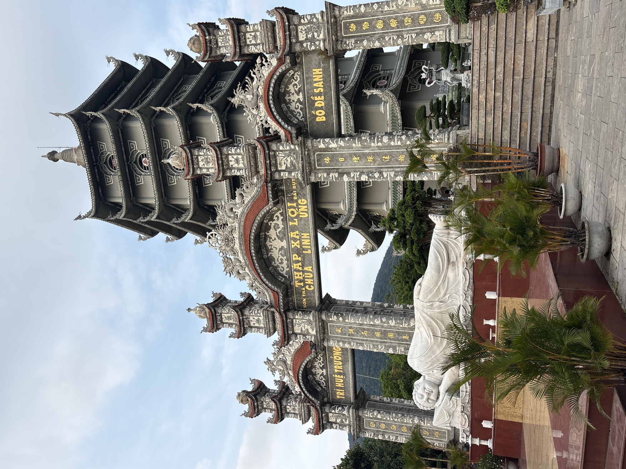 The image shows a vertical view of a stone entrance to a temple complex in Đà Nẵng, Vietnam. Intricate carvings and script in gold lettering adorn the grey-toned structures of the entry gate. The script reads: "TRÍ HUỆ TRƯỞNG," "SON TRA THÁP XÁ LỢI CHÙA LINH ƯNG," and "BỒ ĐỀ SANH." A reclining white statue of a Buddha lies on the right side of the gate, positioned on a red platform decorated with greenery. A flight of stairs can be seen through the gate, leading further into the temple complex. In the background, a mountain is visible through the gate, and the sky is partly cloudy.