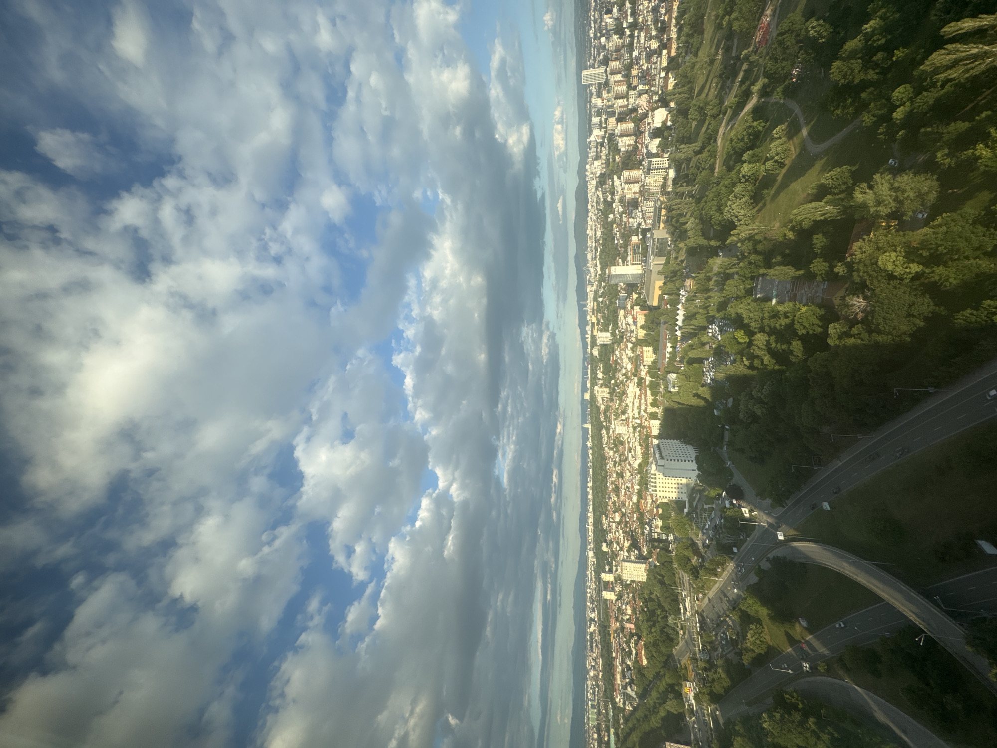 This is an aerial, high-angle, full shot of a city skyline at dusk, taken from an elevated position, likely a tall building. The image captures a sprawling urban landscape characterized by a dense collection of residential and commercial buildings, interspersed with areas of lush green parkland. The city is framed by a backdrop of distant mountains under a sky filled with dramatic, volumetric clouds.
The foreground features a complex interchange of multi-lane roads with overpasses and underpasses, demonstrating significant urban infrastructure. Several vehicles are visible on the roads, suggesting moderate traffic flow. The roads wind through a large, verdant park that dominates the mid-ground of the image. This park is characterized by rolling hills, scattered trees, and winding pathways. Some trees are casting long shadows, indicating the late hour of the day.
Beyond the park, the city unfolds with a variety of architectural styles, ranging from low-rise apartment buildings with terracotta roofs to taller, modern white and light-colored buildings, including what appears to be a hotel or office building with many windows. The density of buildings suggests a well-populated urban center. In the distance, the hazy outlines of mountains can be seen, adding a natural element to the urban panorama.
The sky is a prominent feature, with a mix of bright blue patches and large, cumulonimbus clouds that are illuminated by the setting sun. The lighting creates a dynamic contrast, with some parts of the city and park brightly lit, while others are in shadow, enhancing the sense of depth and time of day.
Based on the architectural styles and the presence of mountains in the distance, this scene is highly likely to be Sofia, Bulgaria. The specific location within Sofia appears to be an area with a mix of residential neighborhoods, commercial zones, and significant green spaces, possibly including areas like the South Park (Yuzhen Park) or Boyana, given the proximity to Vitosha Mountain, which is likely represented by the distant mountains. The elevated viewpoint suggests the photograph was taken from a high-rise building in the southwestern part of the city, overlooking the park and the city center towards the northwest. No individuals are clearly discernible in the image due to the distance and resolution.